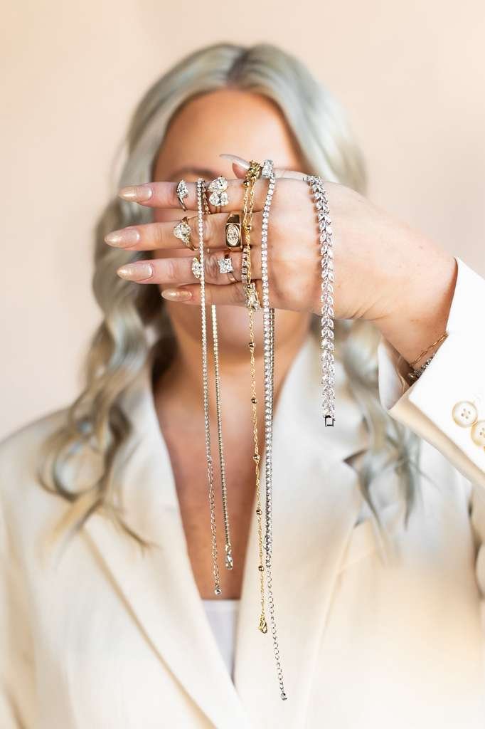 A woman with light gray hair, dressed in a white blazer, holds up her hand showing numerous rings and necklaces, covering her face.