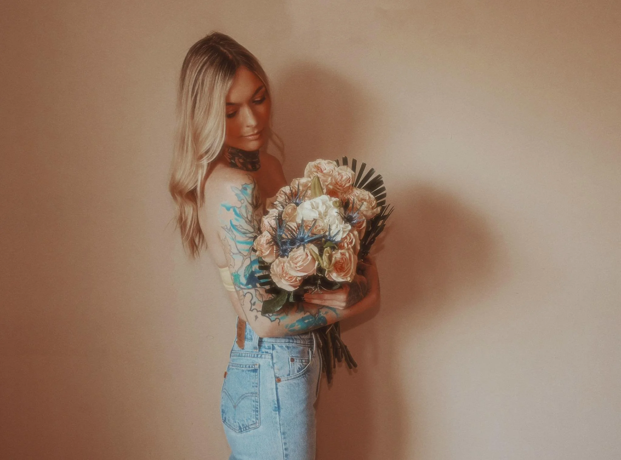 Woman with blonde hair and tattoos holding a bouquet of flowers against a beige wall.