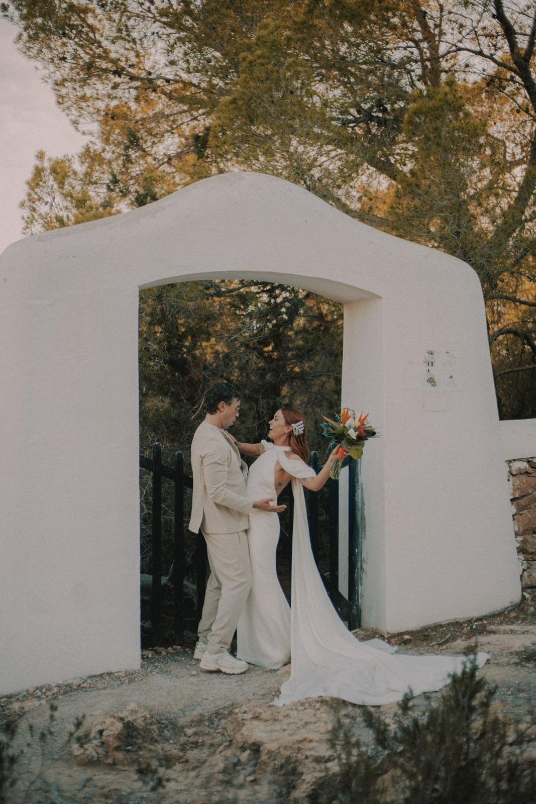 A bride and groom are standing under a white archway outdoors, with trees in the background. The bride is holding a bouquet of flowers and leaning towards the groom, who is embracing her.