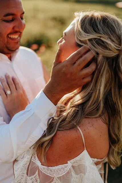 A man gently touching a woman's face outdoors during sunset, both smiling and looking at each other.