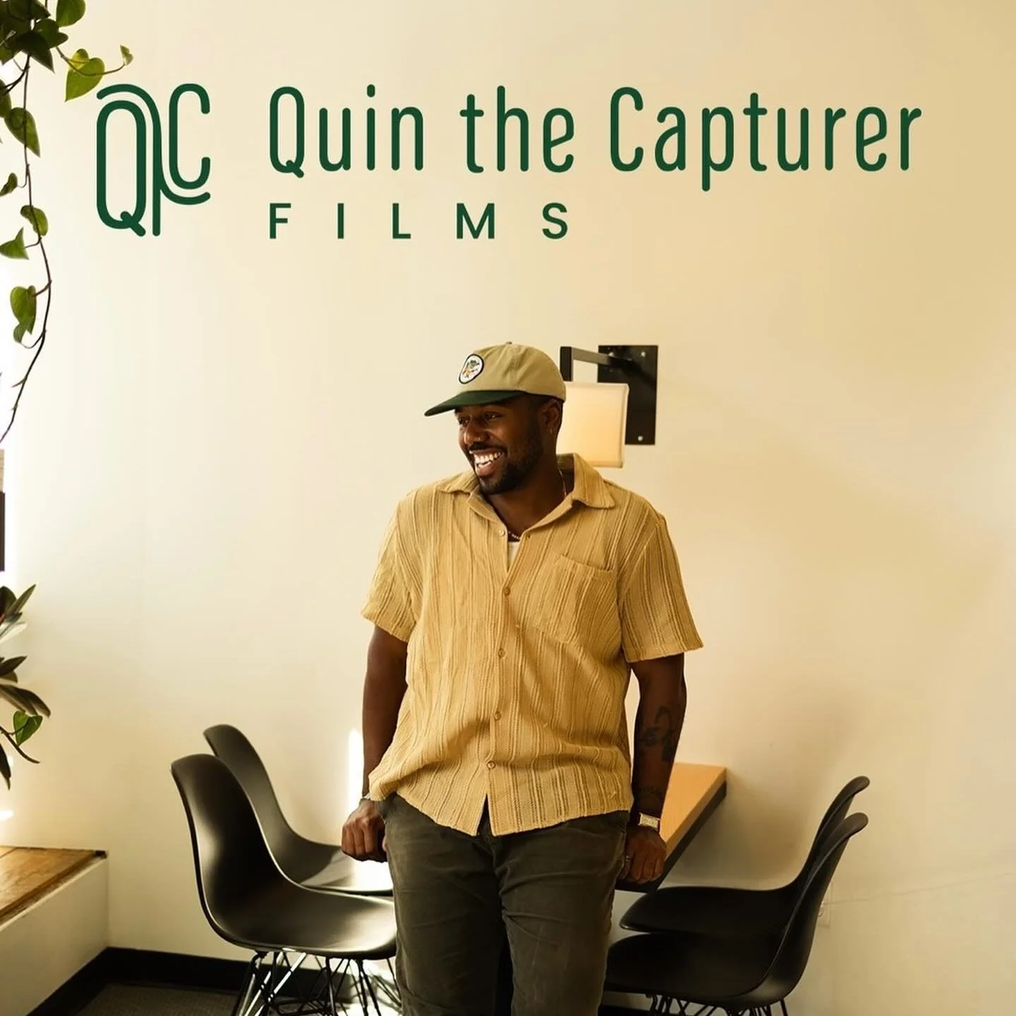 Man in beige cap and yellow shirt smiling and leaning against a table in front of a wall with 'QC Quin the Capture FILMS' sign.