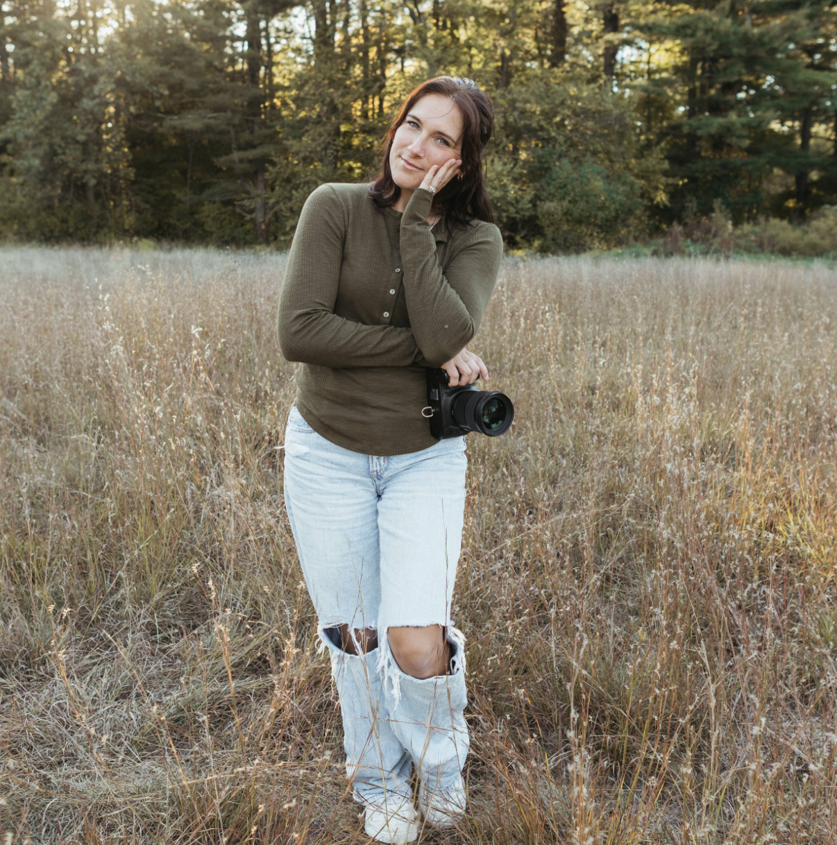 A woman standing in a field during fall, holding a camera, dressed in a green long-sleeve shirt and ripped light jeans, with autumn trees in the background.