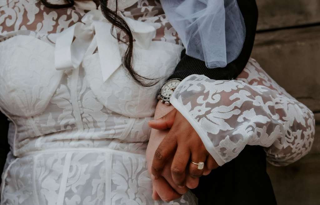 Close-up of a woman wearing a white lace dress with puffed sleeves and a bow at the neckline, holding hands with a person wearing a dark suit, with a wooden background visible.