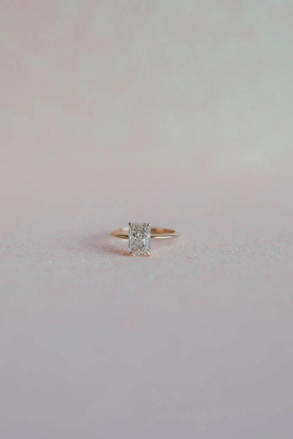 A gold ring with a rectangular diamond solitaire on a soft pink background.