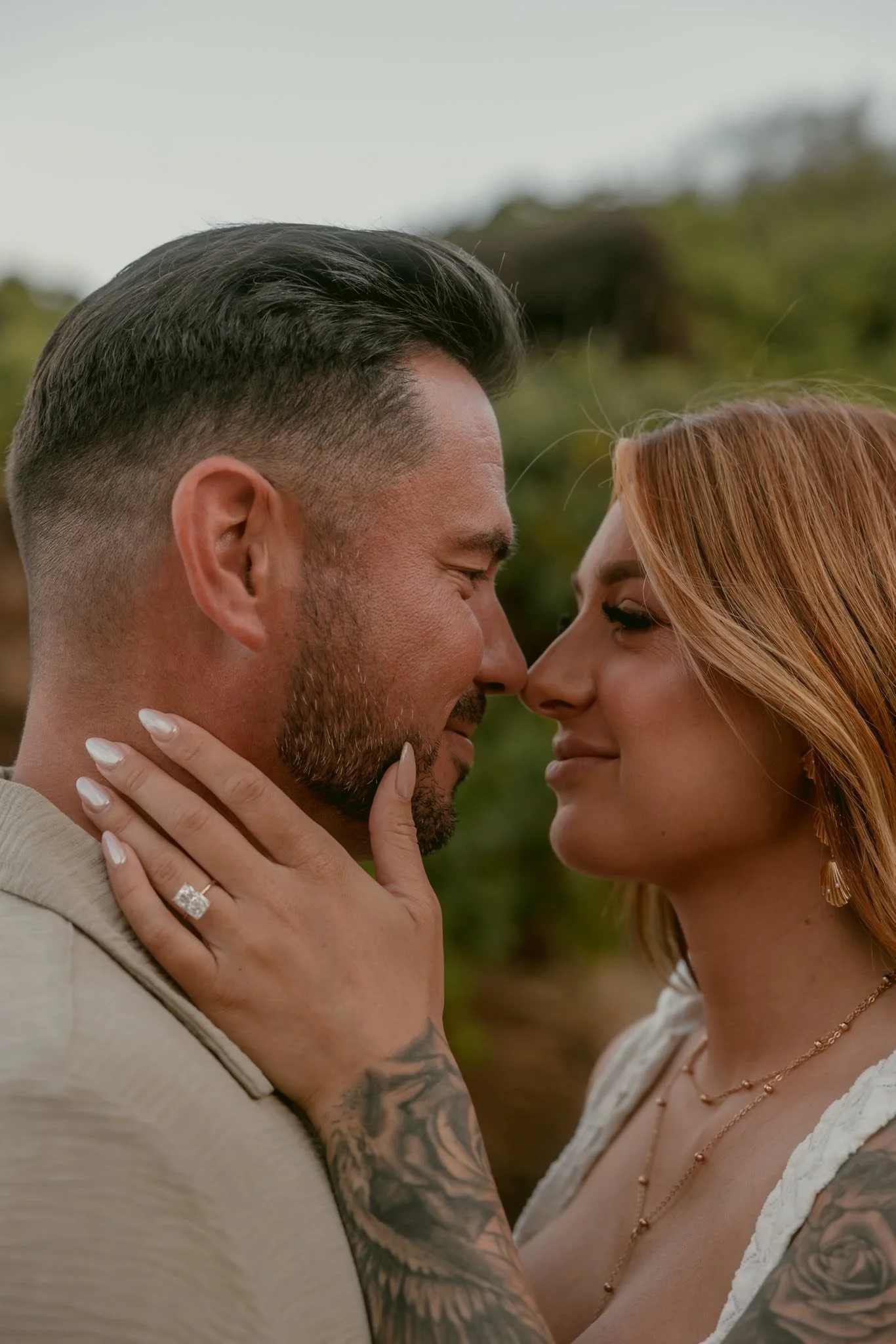 A couple is close together, nose to nose, with the woman gently holding the man's jawline. The woman has red hair, and tattoos on her arm and chest. The man has short dark hair and a beard. They are outdoors with a blurred green background.