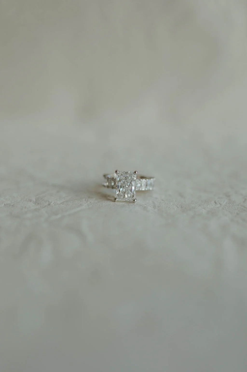A diamond engagement ring with a large rectangular center stone and smaller side stones on a light, textured surface.