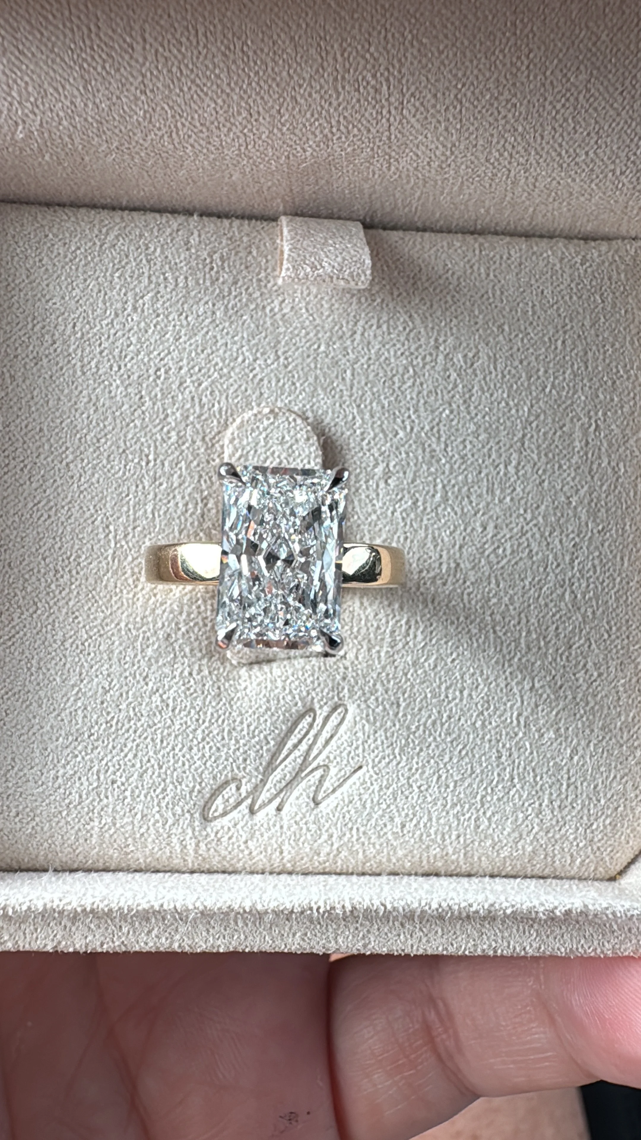 A large rectangular diamond engagement ring with a gold band inside a beige ring box with the initials 'dh' embossed on it.