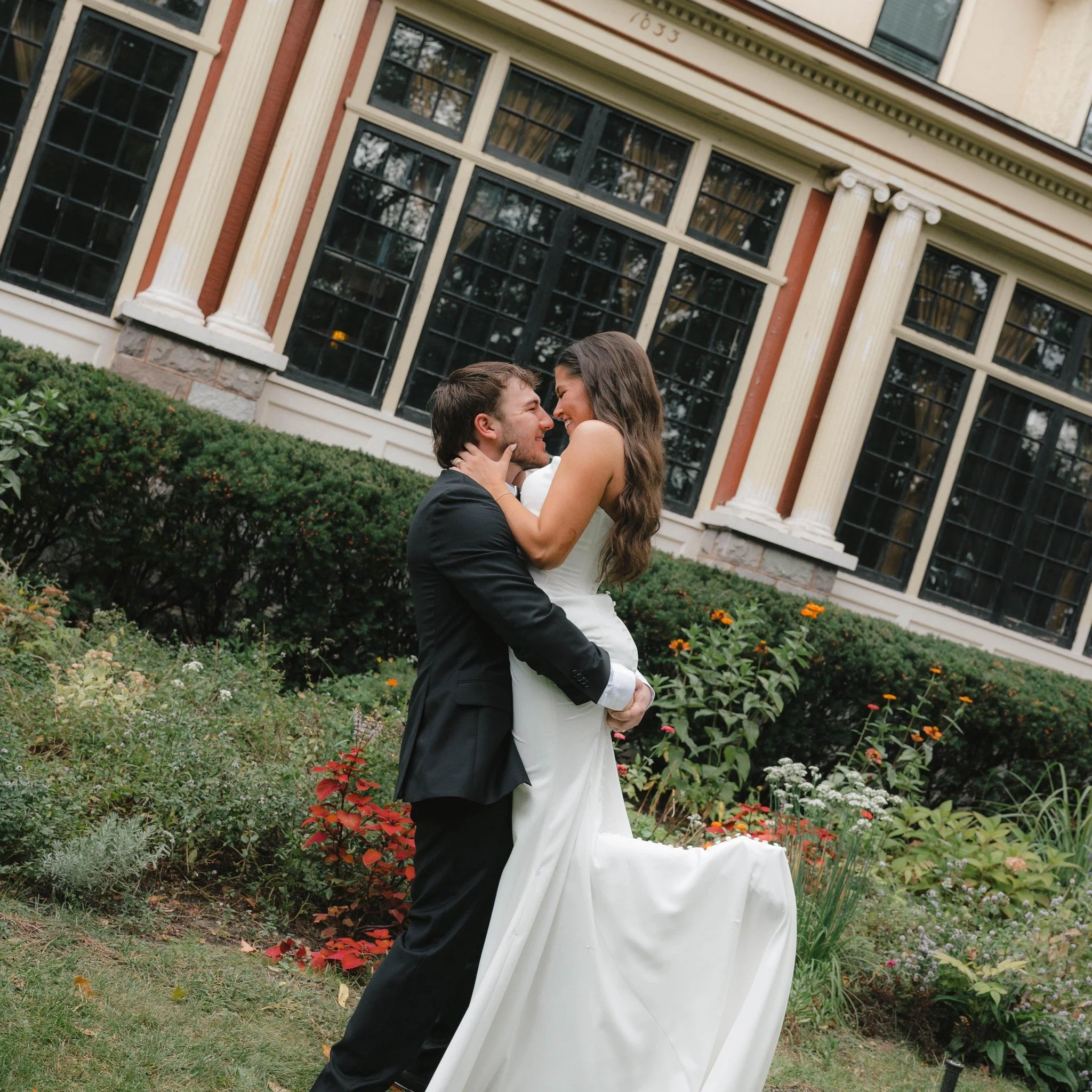 A newlywed couple in wedding attire embracing and smiling in a garden with a large historic building in the background.