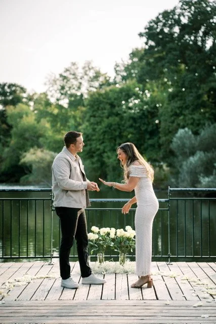 A couple stands holding hands on a wooden platform near a railing with a river and lush green trees in the background, during what appears to be a wedding or special occasion.