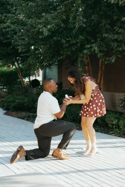 A man is kneeling on one knee, proposing to a woman outdoors near a tree, with a background of greenery and a building.