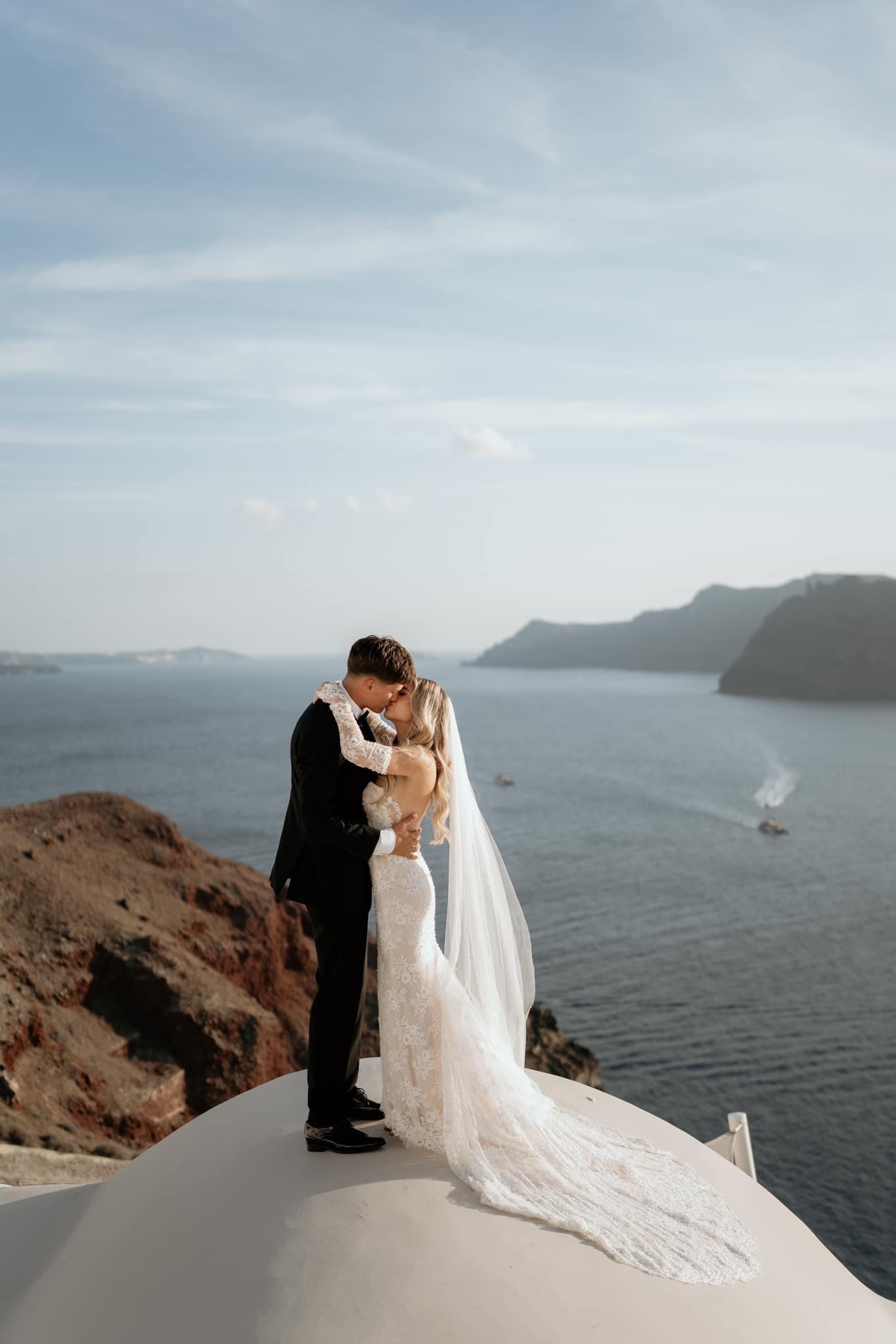Bride and groom sharing a kiss on a hilltop overlooking the sea, with cliffs and boats in the background.