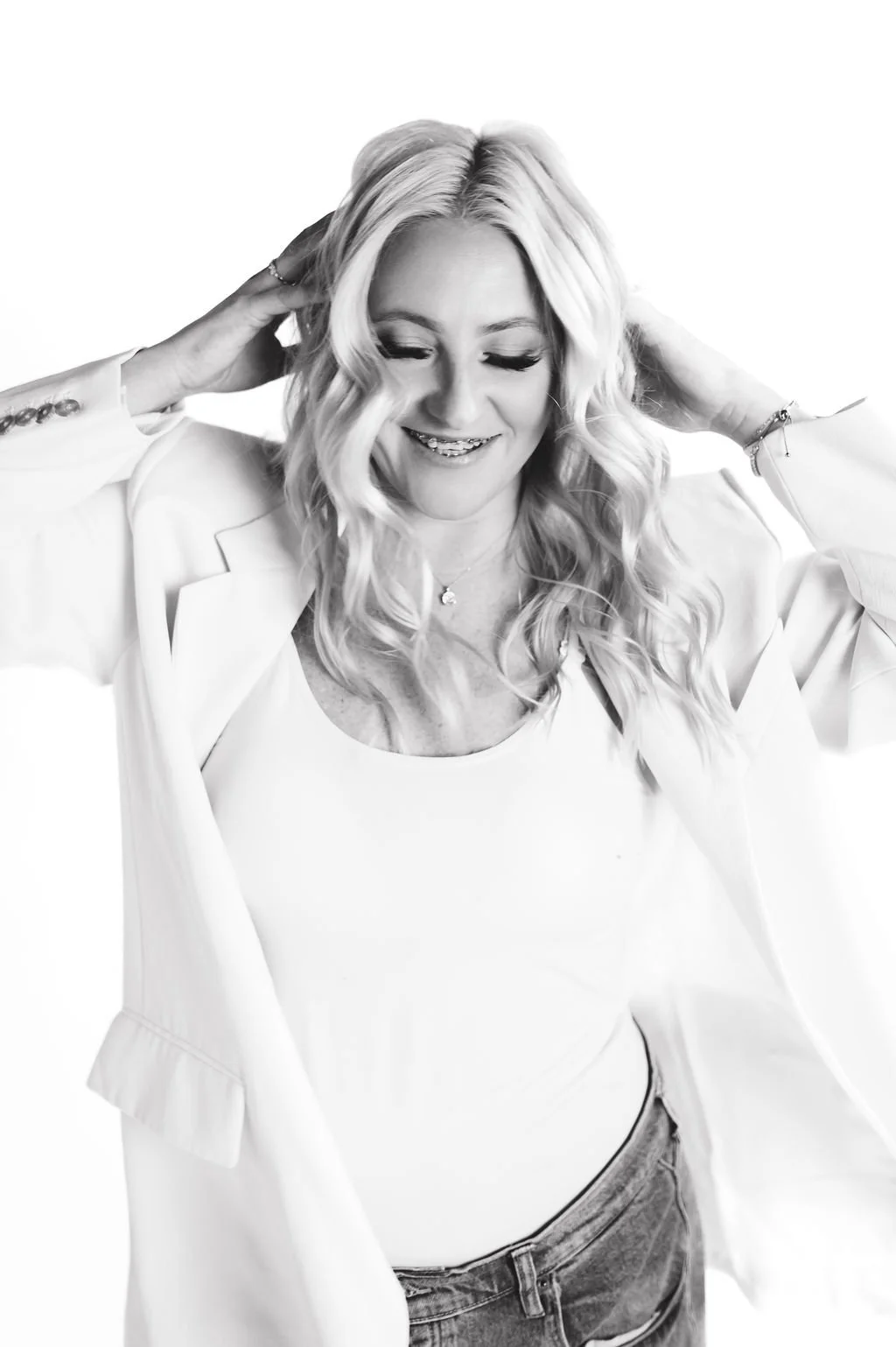 A smiling woman with long, wavy blonde hair wearing a white blazer over a white top and jeans, with her hands behind her head against a plain white background.
