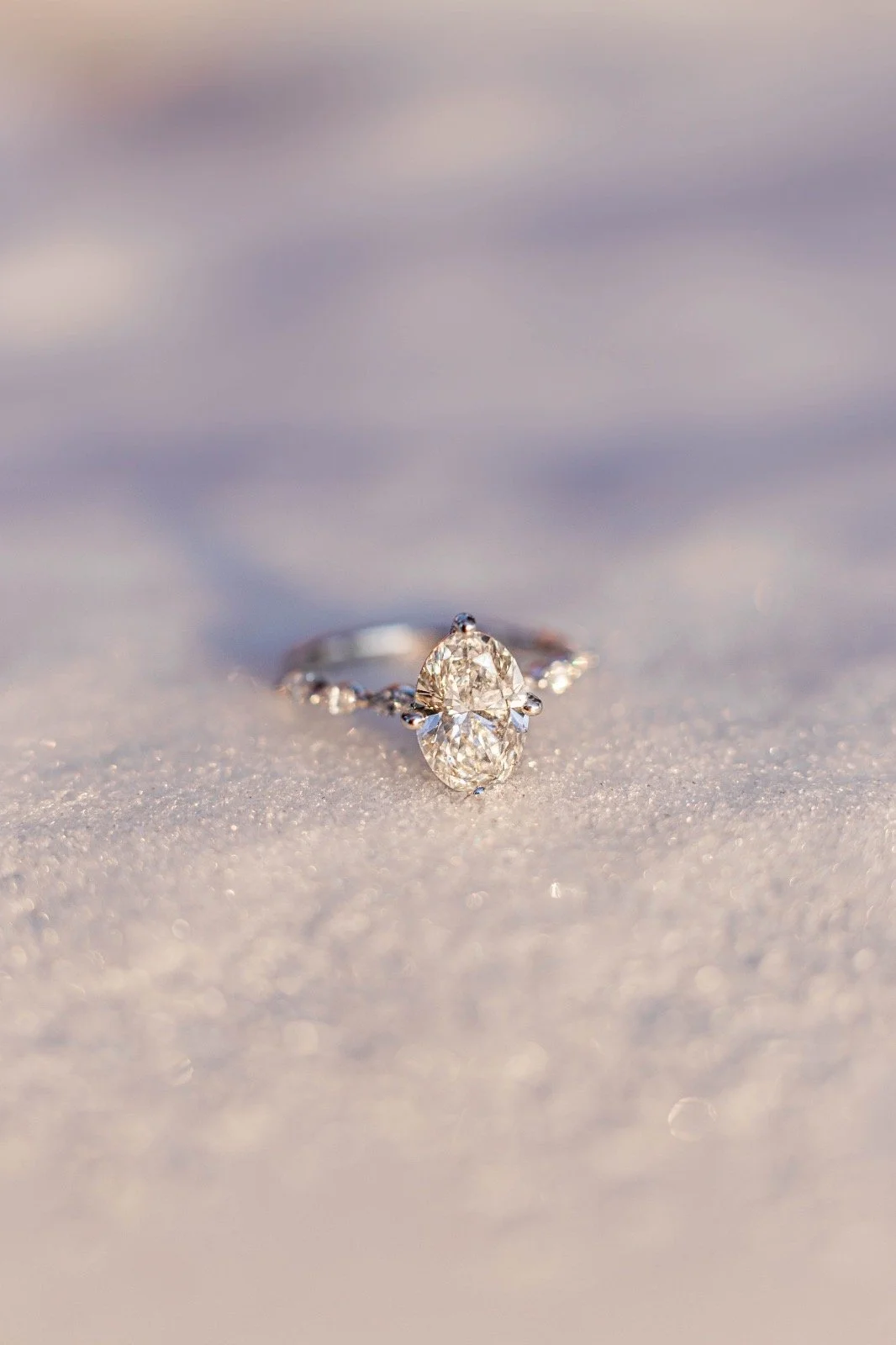 A diamond engagement ring with an oval diamond setting on a sandy surface.