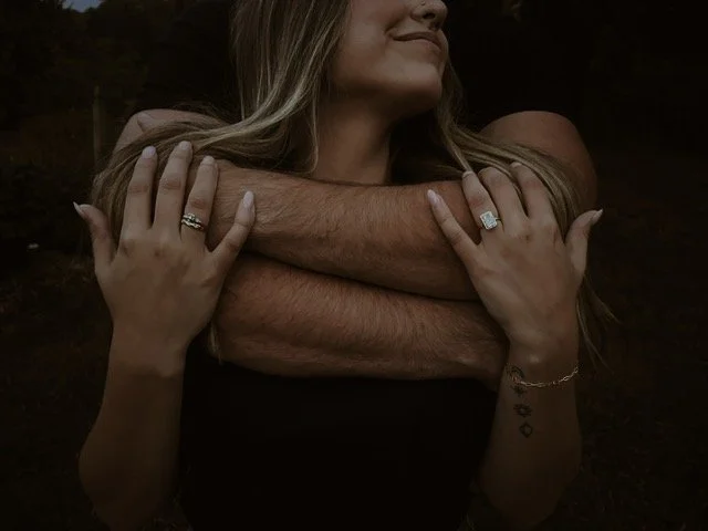 A woman receiving a hug from a man with his arms around her shoulders, both showing rings on their hands.