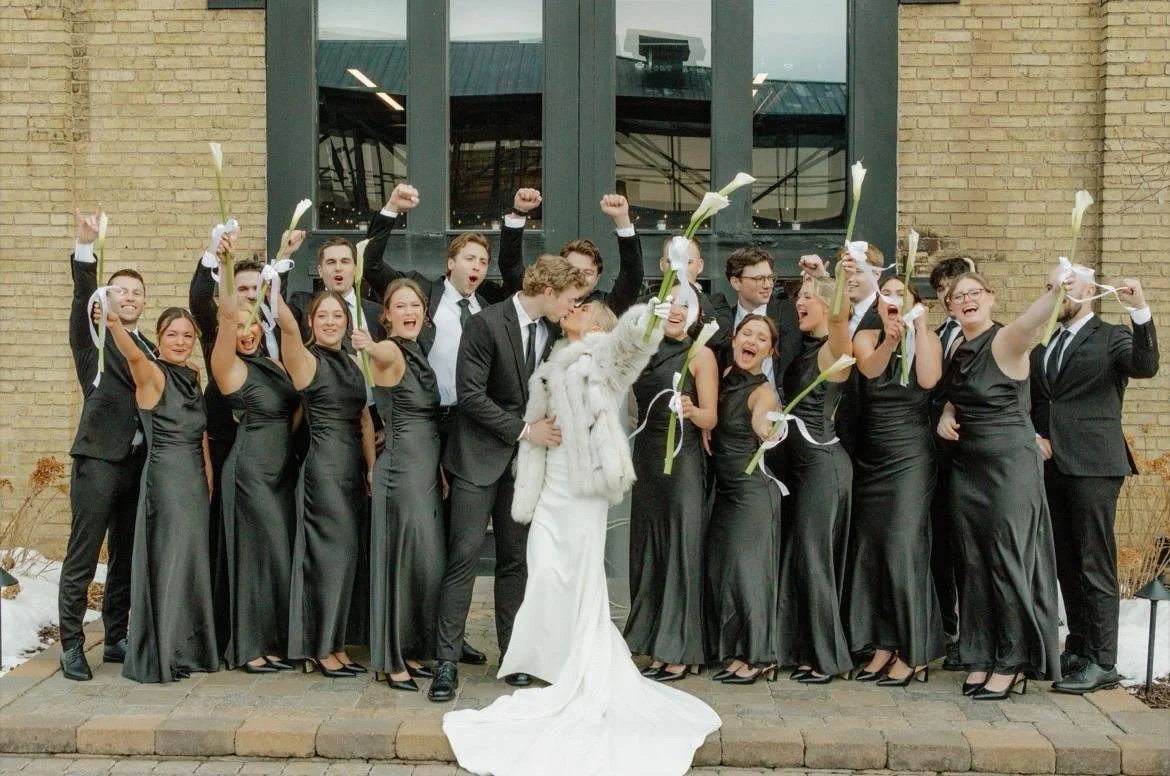 A wedding celebration with the bride and groom kissing in front of a group of people, who are cheering and holding calla lilies, outside a building with brick walls and large windows.