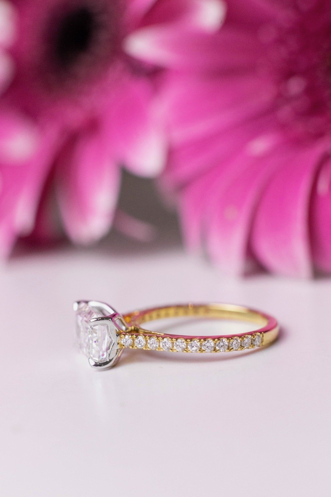 An elegant gold ring with a large central diamond and smaller surrounding diamonds, placed on a soft, pale surface with pink flowers in the background.