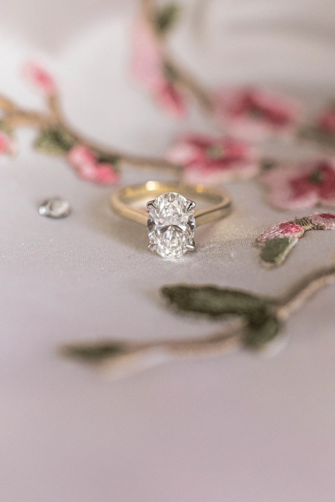 Close-up of a diamond engagement ring with a large oval-cut diamond, surrounded by flowering branch and small gemstones on soft fabric background.