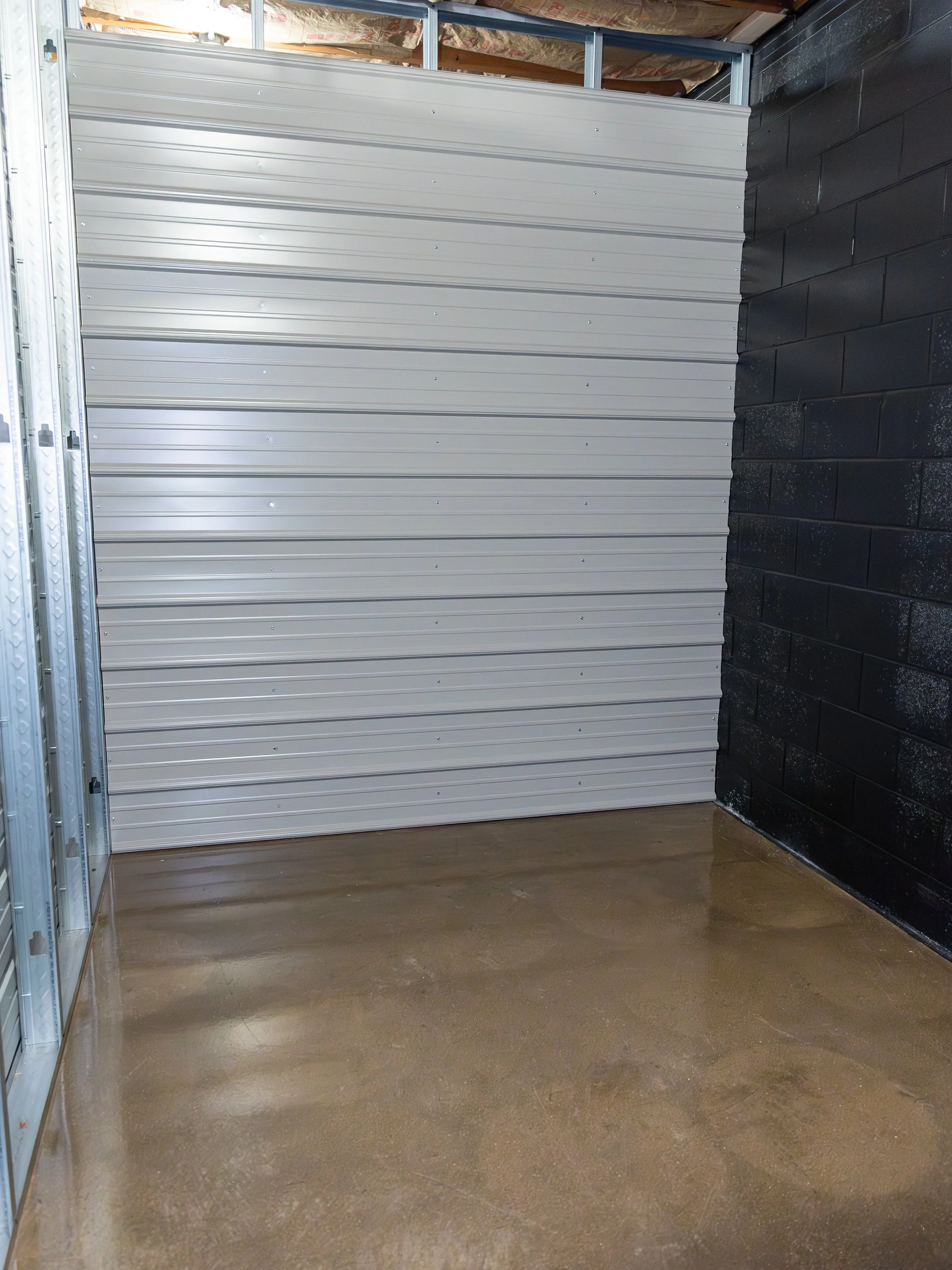 Empty garage with a metal rolling door, black painted brick wall, and a concrete floor.