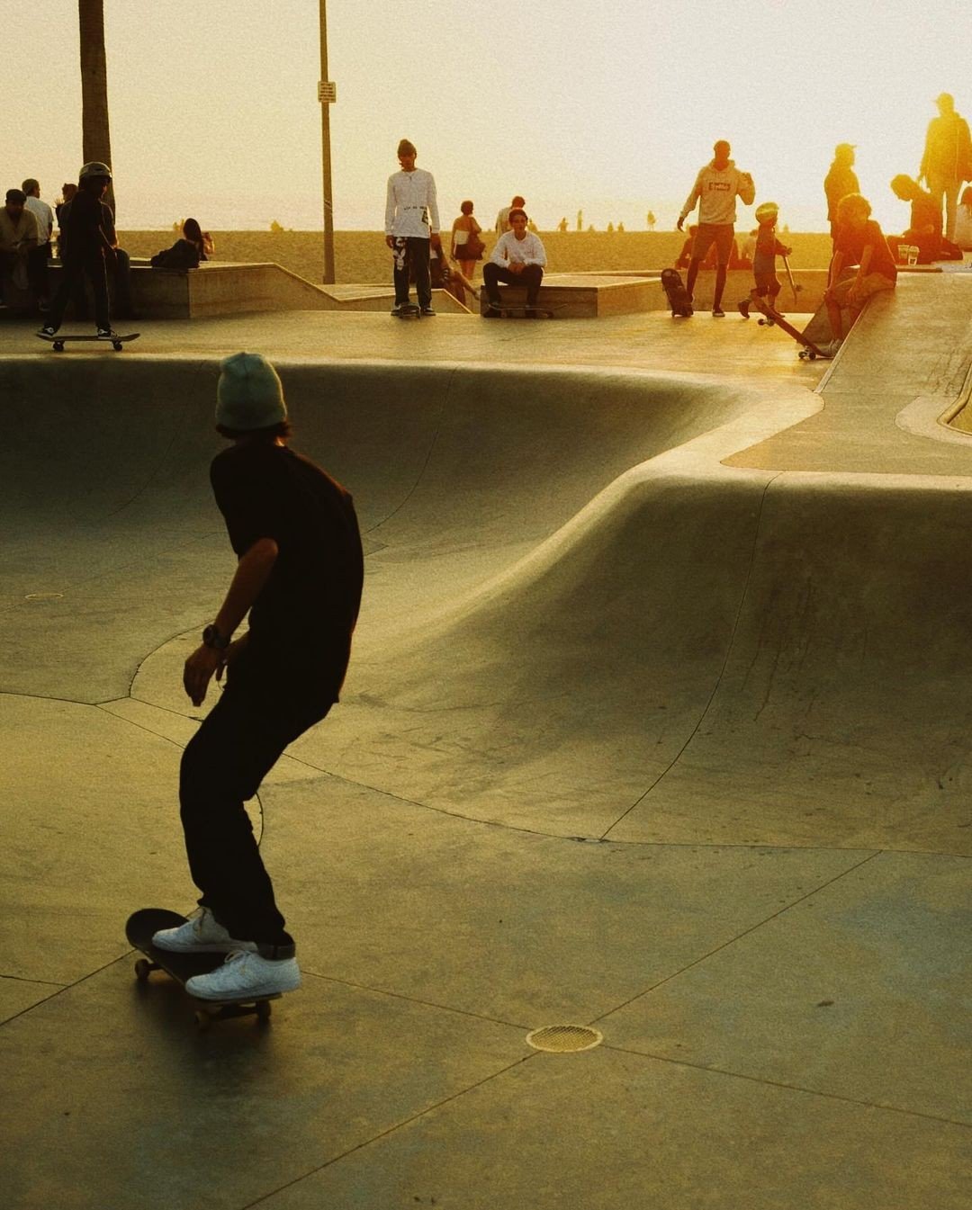 A skateboarder in a black outfit and white shoes glides across a skate park with a bowl and ramp. Several other skaters and spectators are present, some sitting and some standing, with a sunset or sunrise sky in the background.
