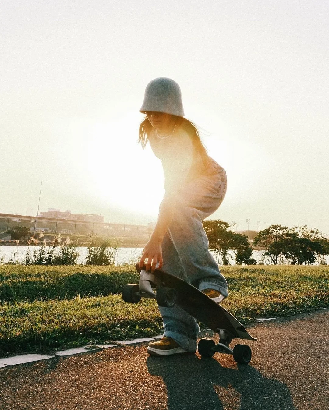 A young woman in casual attire, including a gray beanie and jeans, is holding a skateboard while squatting on a pathway near a body of water during sunset.