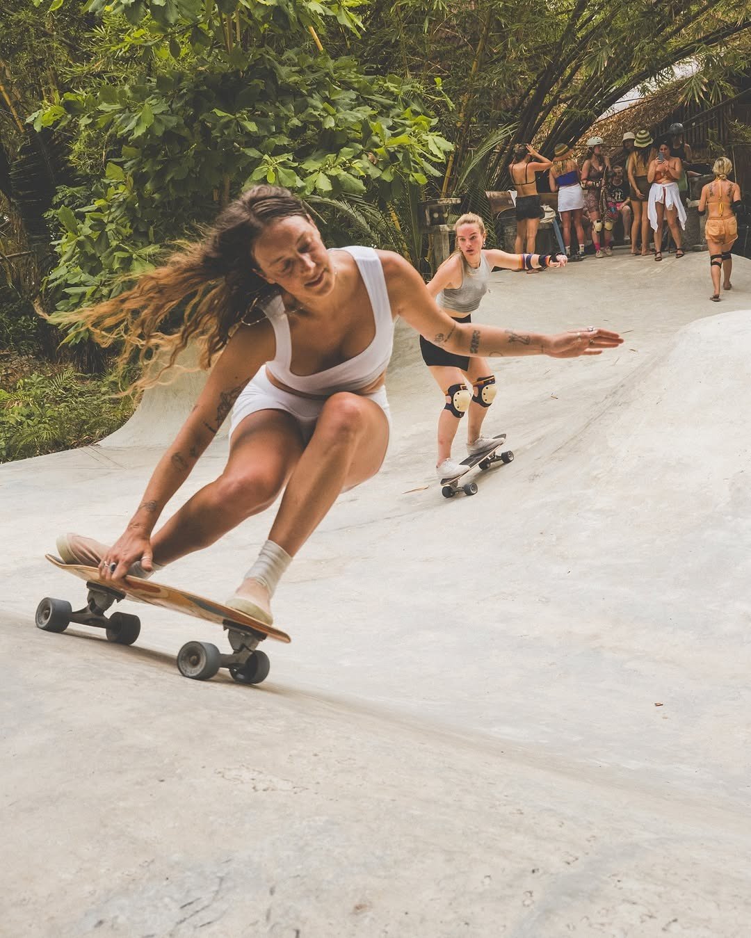 Two women skateboarding on a concrete skate park while a group of people watches in the background.