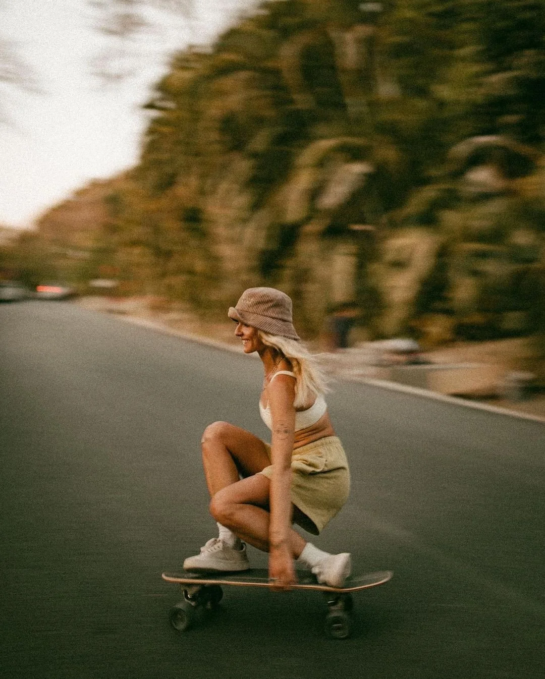 A woman skateboarding on a road with trees in the background, wearing a bucket hat, white tank top, beige shorts, and sneakers, smiling as she rides.