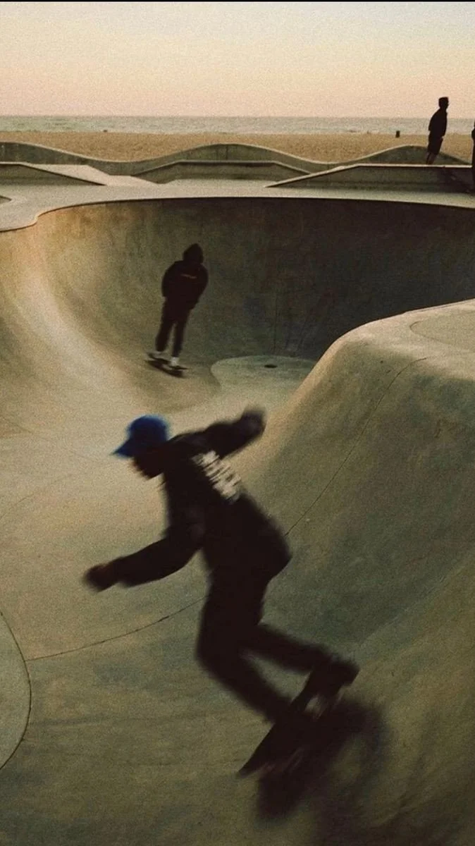 People skating at a concrete skatepark during dusk, with the ocean in the background.