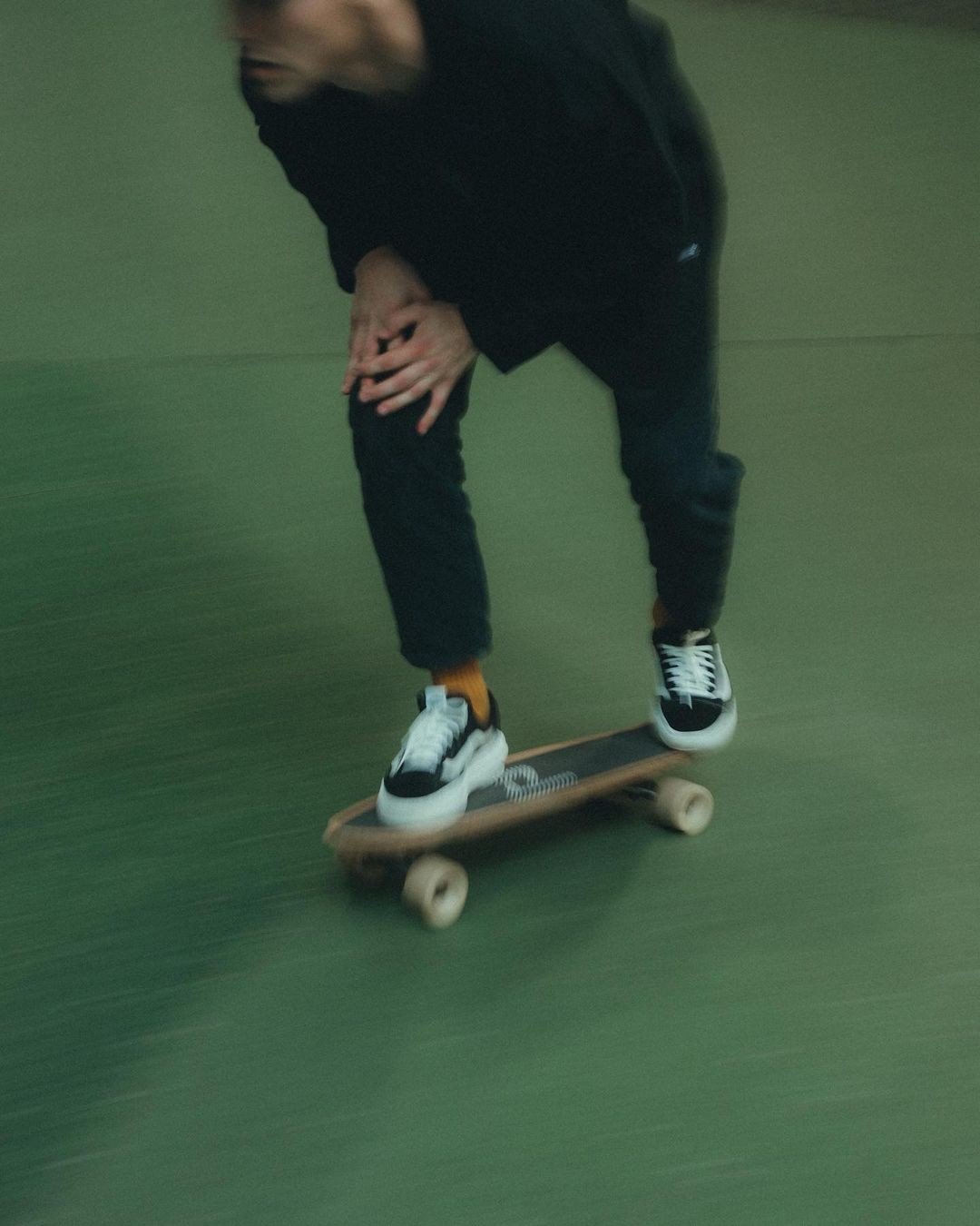Person riding a skateboard indoors, wearing black clothes and white sneakers, with one hand on their knee, slightly bent forward.