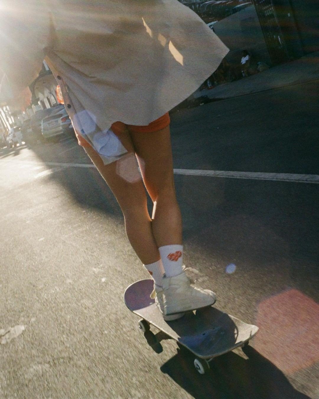 Person skateboarding on a city street during sunset, wearing a white shirt, orange shorts, white shoes, and white socks with orange logo.