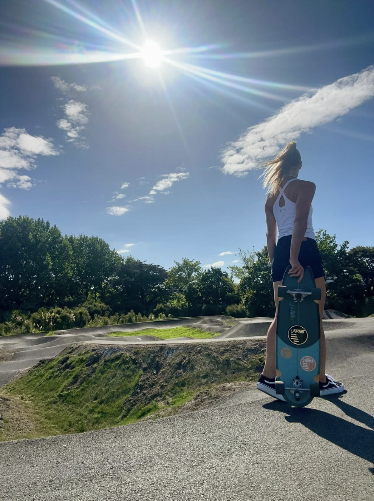 A woman standing on a skate park, holding a skateboard, with a bright sun in the sky and trees in the background.