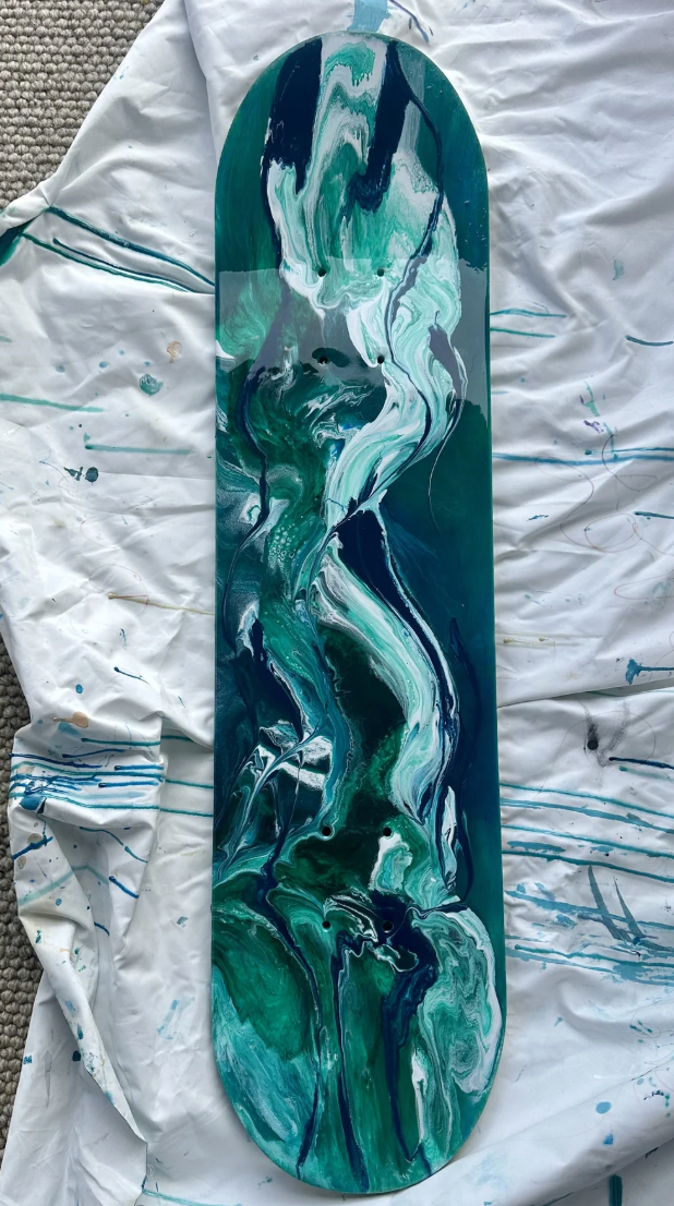 An artist's skateboard deck featuring an abstract swirling design in shades of teal, blue, white, and black, placed on a paint-splattered white cloth.