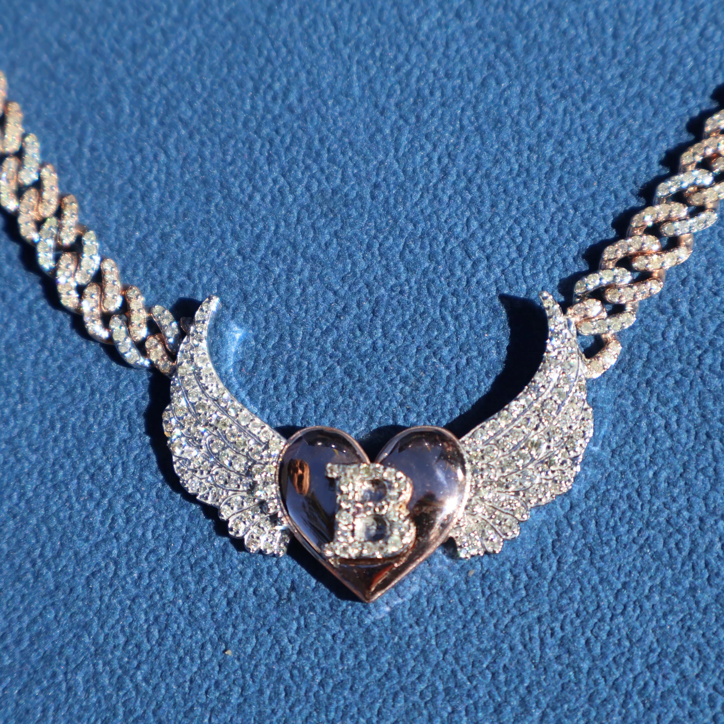 A white gold necklace with large diamond-encrusted wings and a heart-shaped pendant featuring the letter B and a small heart inside on a blue textured surface.