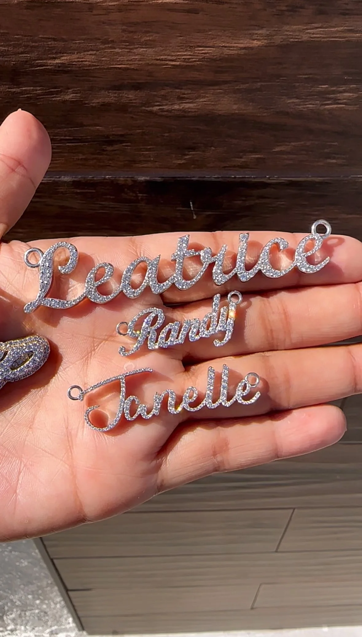 Hand holding three silver and diamond custom nameplate jewelry pieces with the names 'Leatrice,' 'Randy,' and 'Janelle' over a wooden table surface.