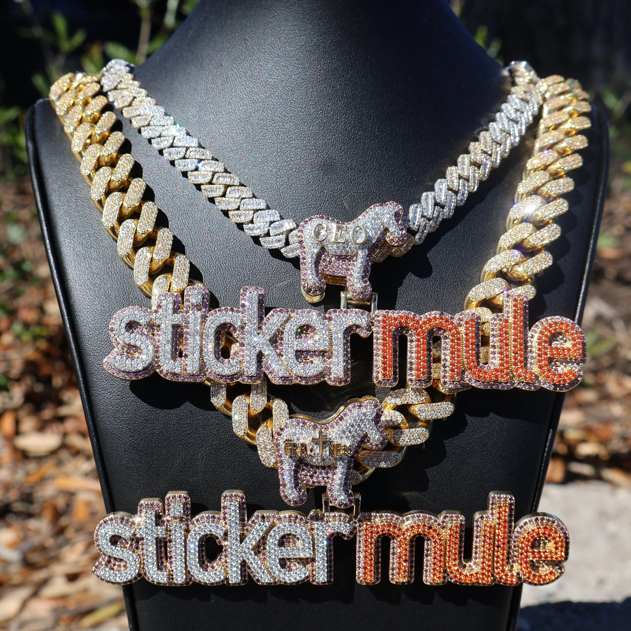 Three large gold and diamond-encrusted necklaces with pendants, displayed on a black jewelry bust. The necklaces feature the words 'sticker mule' in rainbow-colored letters and small horse-shaped charms.