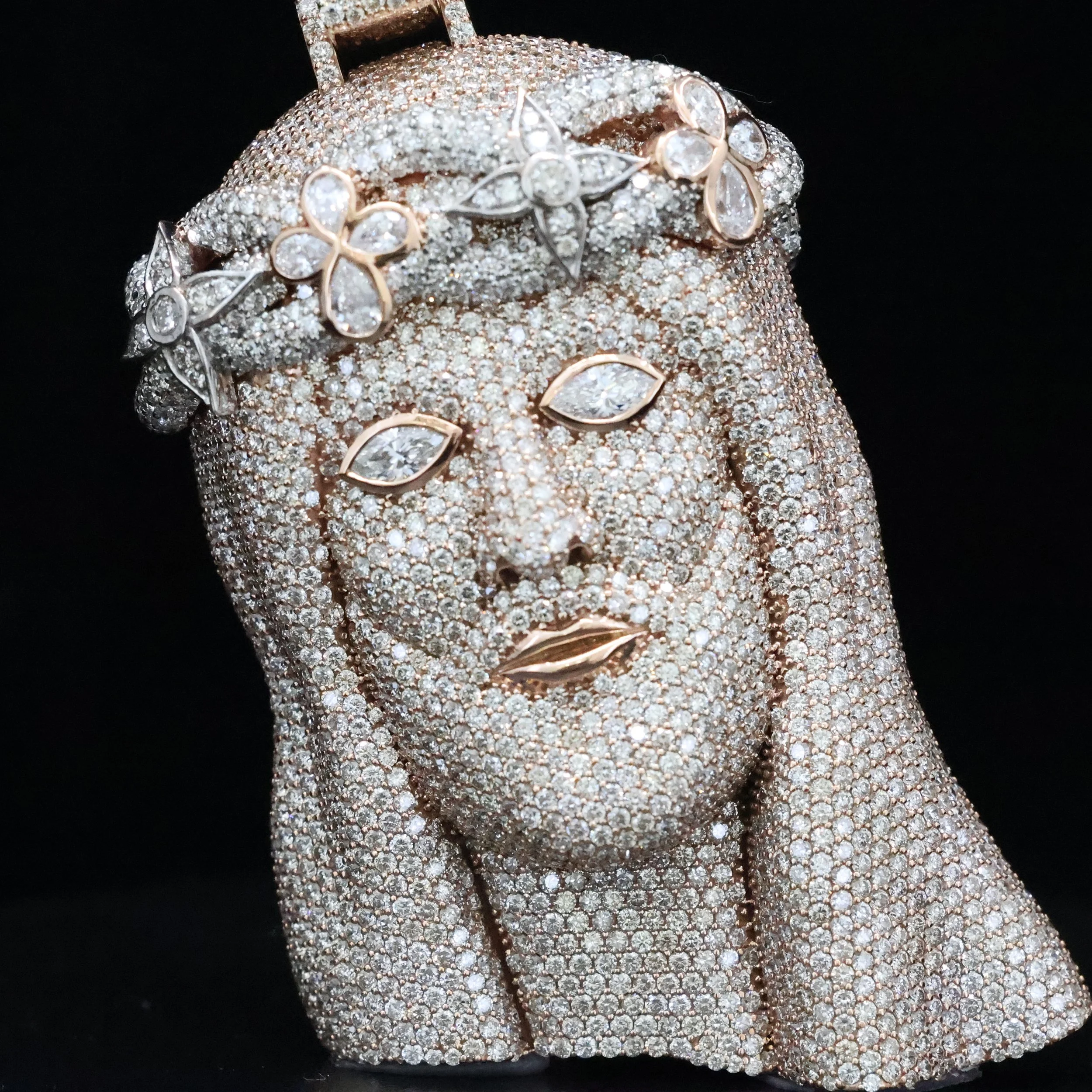 A custom jewelry piece in the shape of a Jesus's face and upper torso, fully covered in small diamonds with gold accents for the lips, eyes, and a floral headpiece, set against a black background.