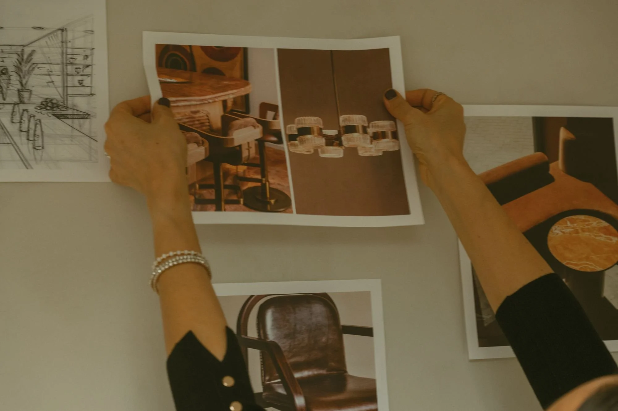 Person holding interior design photographs, one showing a bar with chairs, another a chandelier, and others of furniture, on a wall.