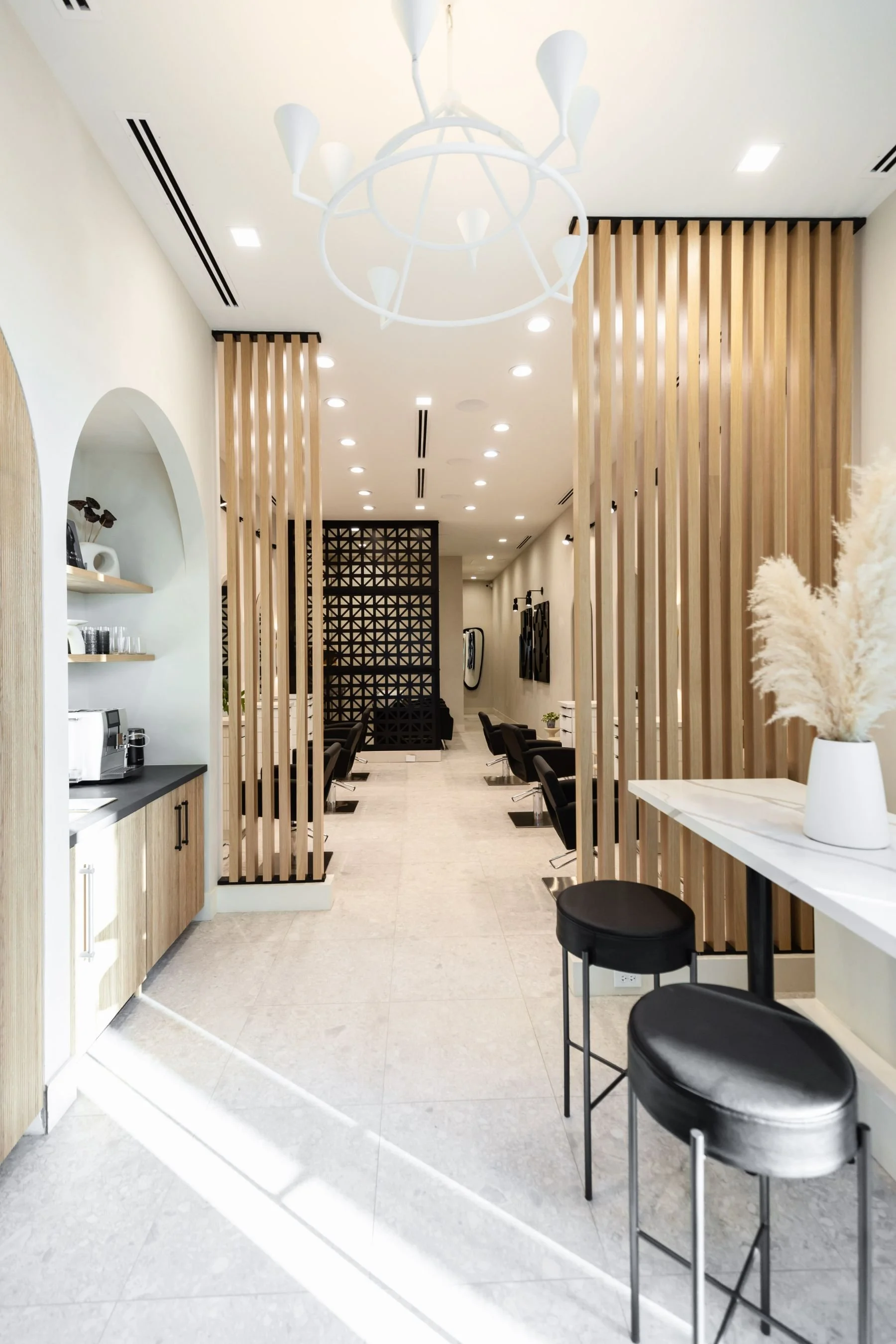 Modern hair salon interior with black chairs, wooden slat dividers, and reception counter with pampas grass arrangement.