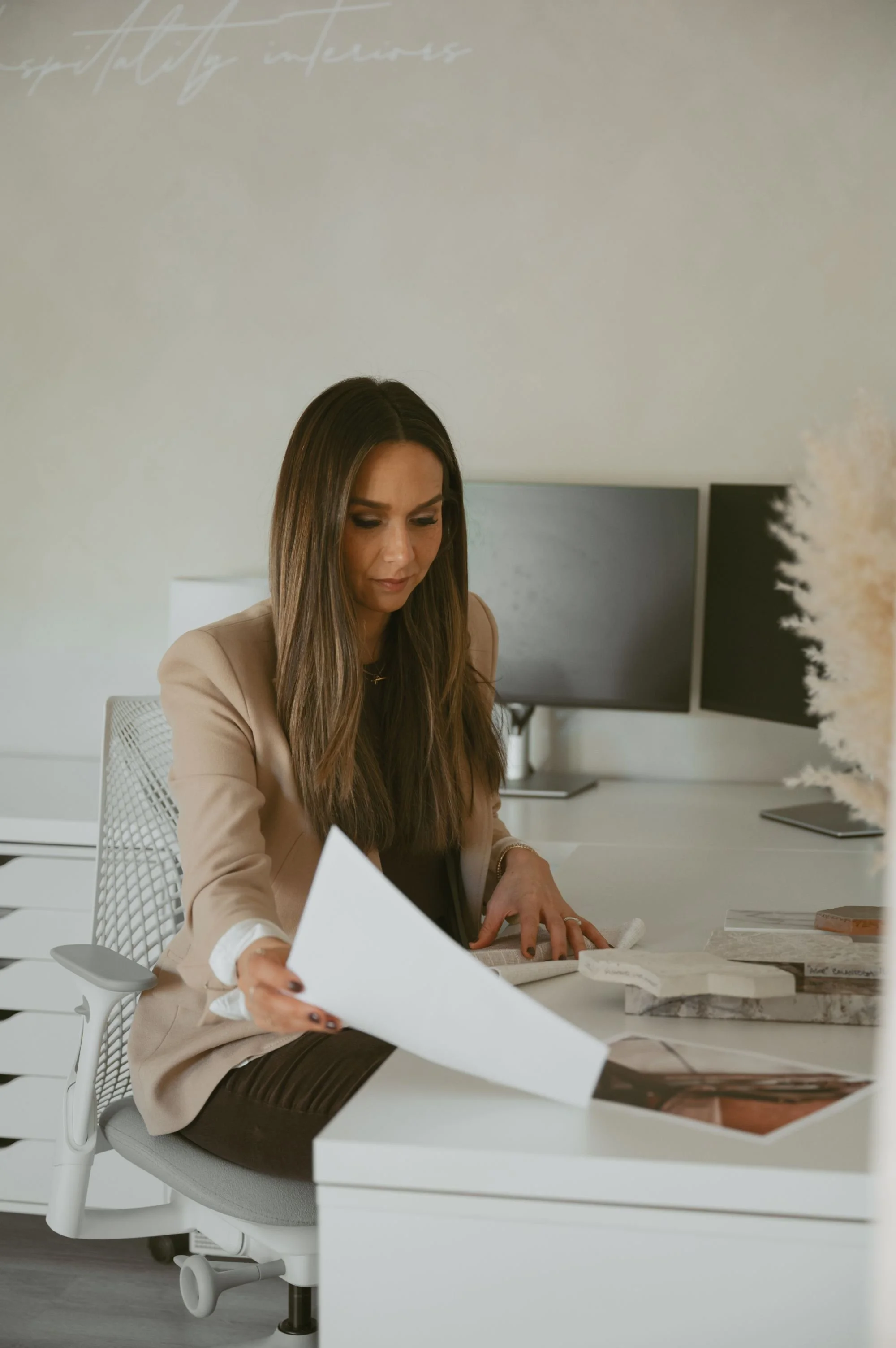 A woman sitting at a white desk, holding a document, with two computer monitors in the background, in an office setting.