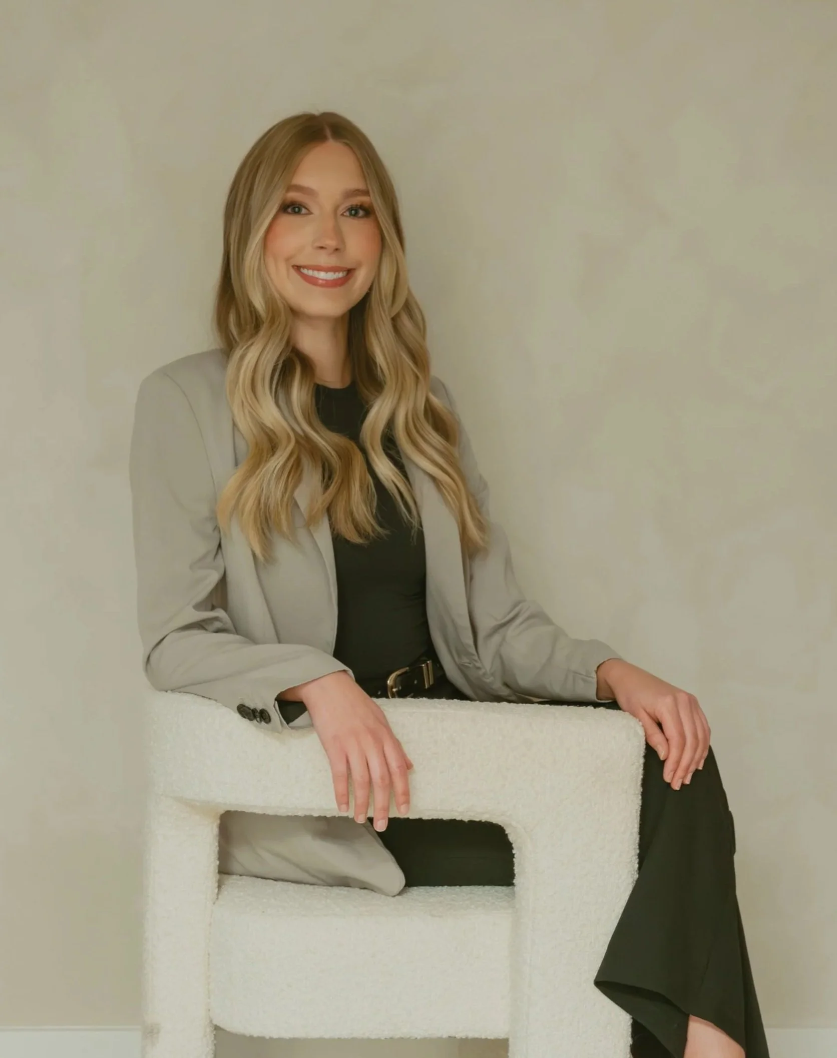 A smiling woman with long blonde hair sitting on a textured white chair, wearing a grey blazer over a black top, in front of a plain light-colored wall.