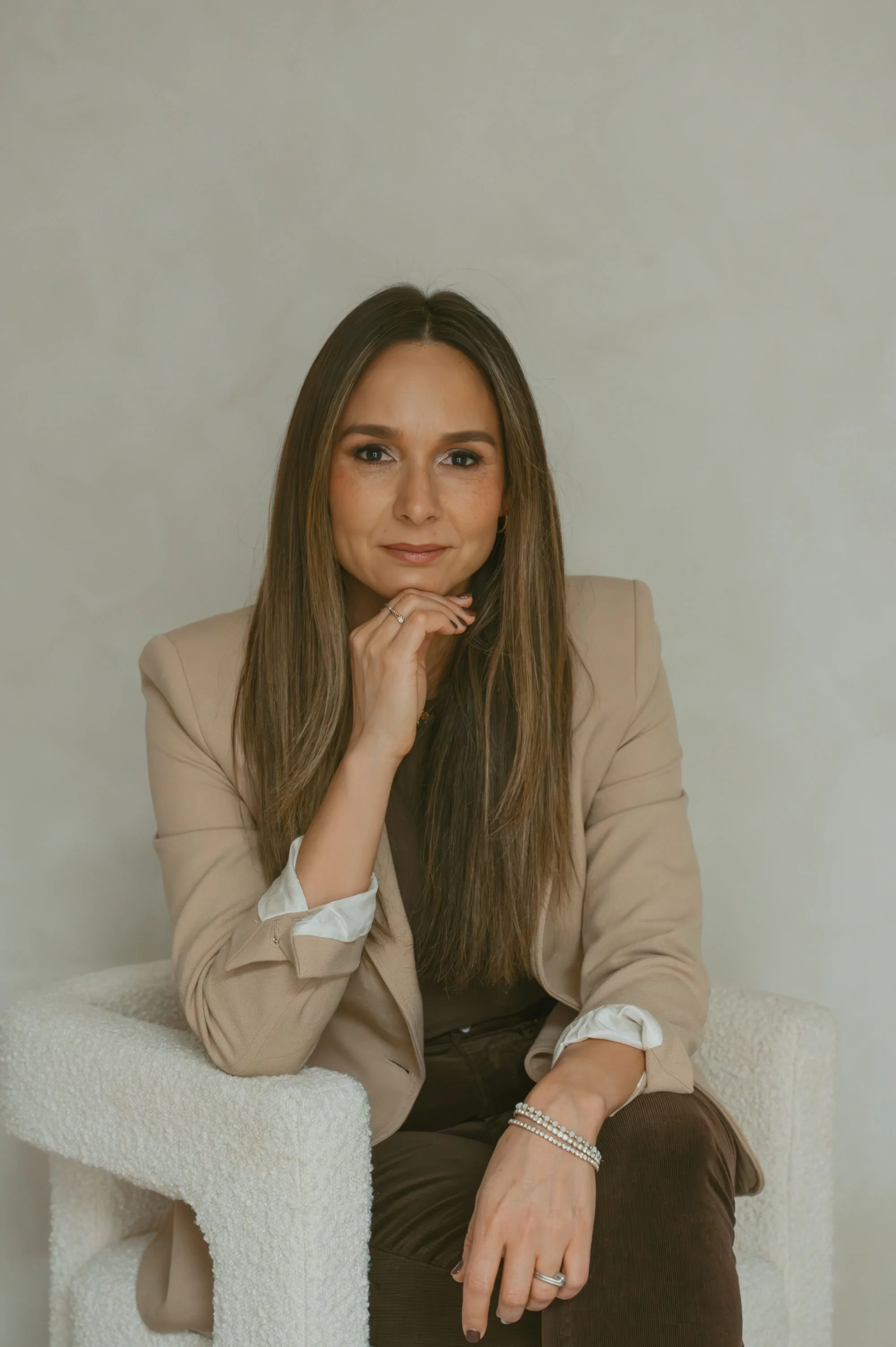 A woman with long brown hair and a slight smile, wearing a beige blazer and brown pants, sitting on a textured white chair against a plain light-colored wall.
