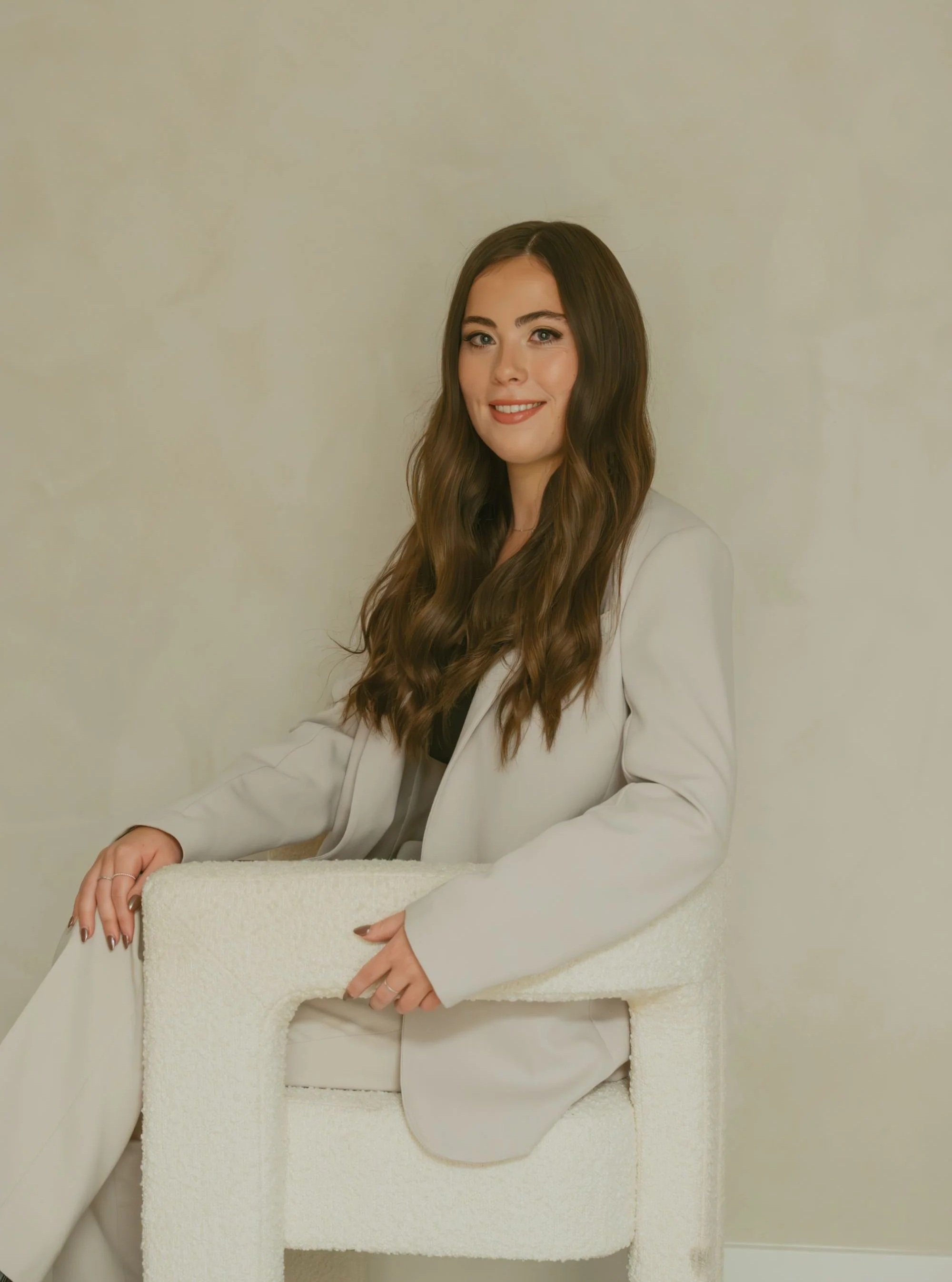 A woman with long, wavy brown hair sitting on a white textured chair, wearing a light-colored blazer and smiling at the camera.
