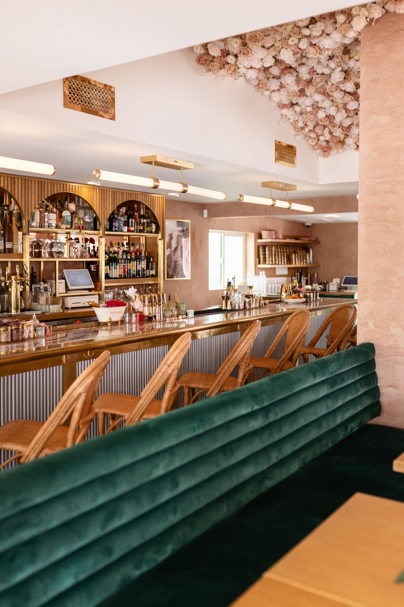 Interior of a stylish bar with a green velvet booth in the foreground, a wooden bar counter with barstools, and shelves stocked with bottles of liquor in the background. Decorative pink flowers are on the ceiling.