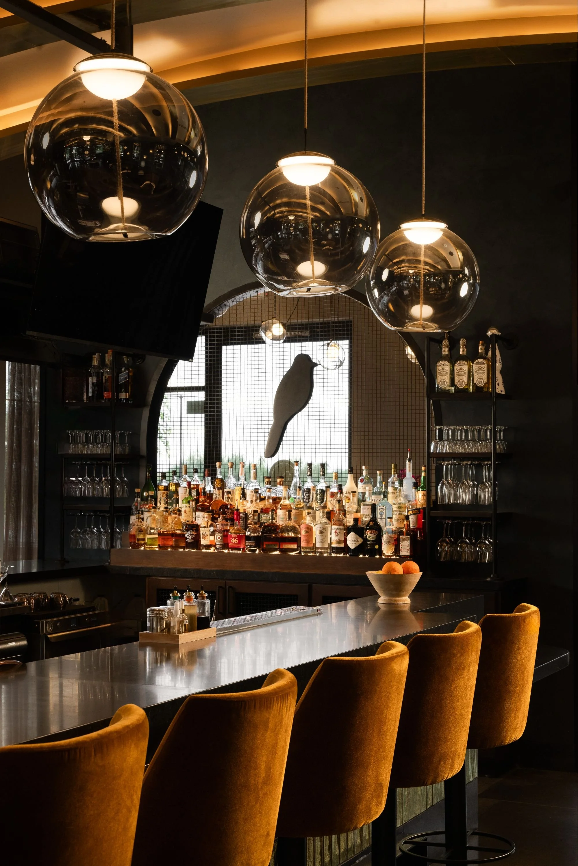 A stylish bar with amber velvet chairs at the counter, modern pendant lights, bottles of alcohol behind the bar, a bowl of fruit, and a decorative hummingbird sculpture on the back wall.