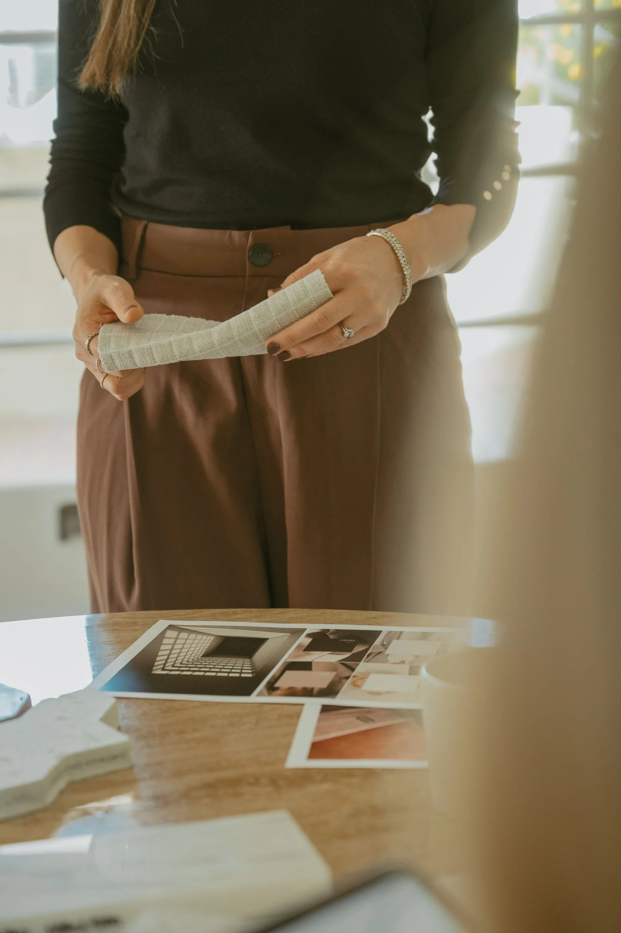 A woman wearing a black long-sleeve top and brown pants is wrapping a bandage around her hand, standing in front of a table with printed photographs and various objects.