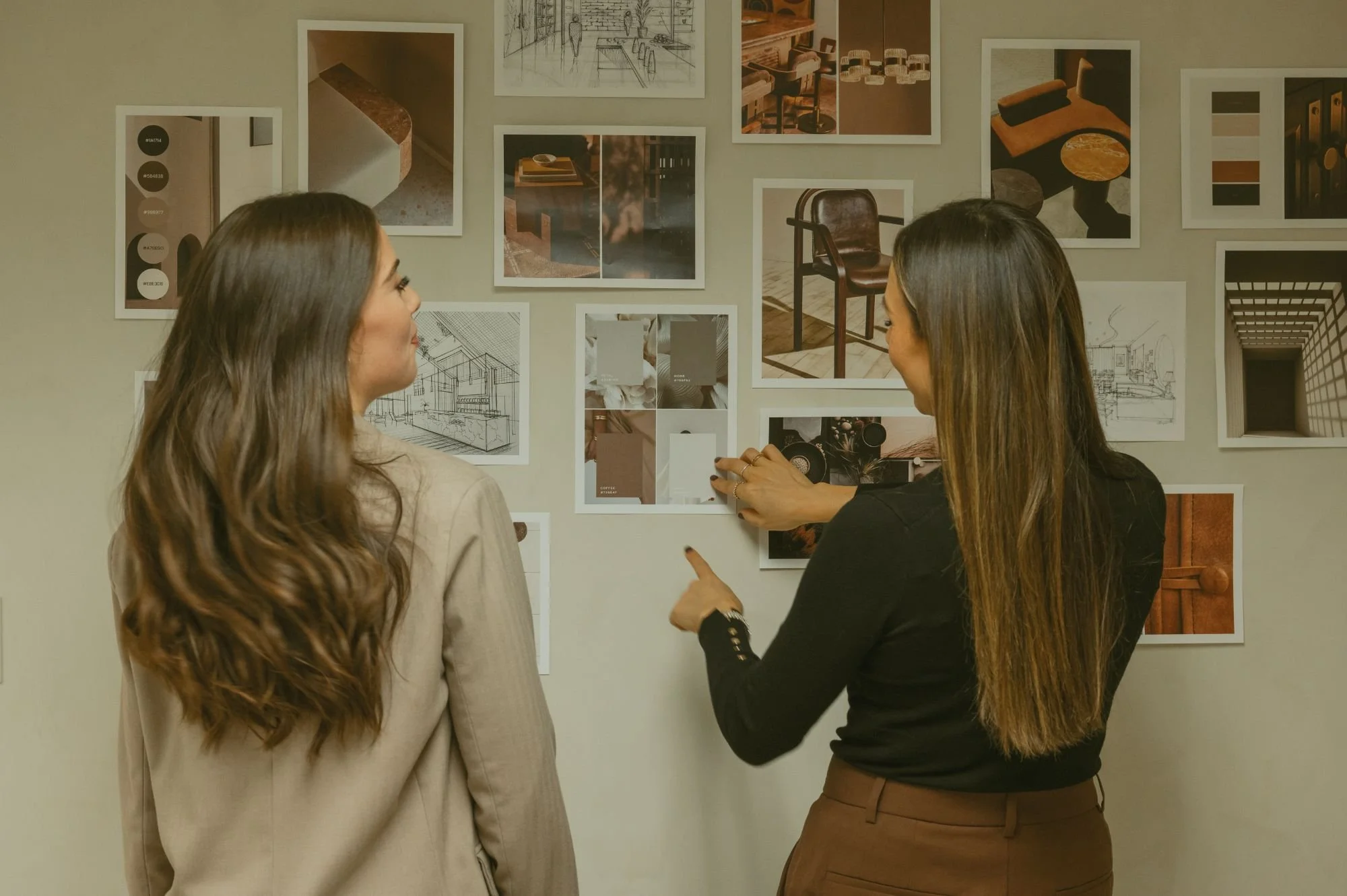 Two women discuss interior design ideas while looking at a wall filled with photos and sketches of room layouts and furniture.