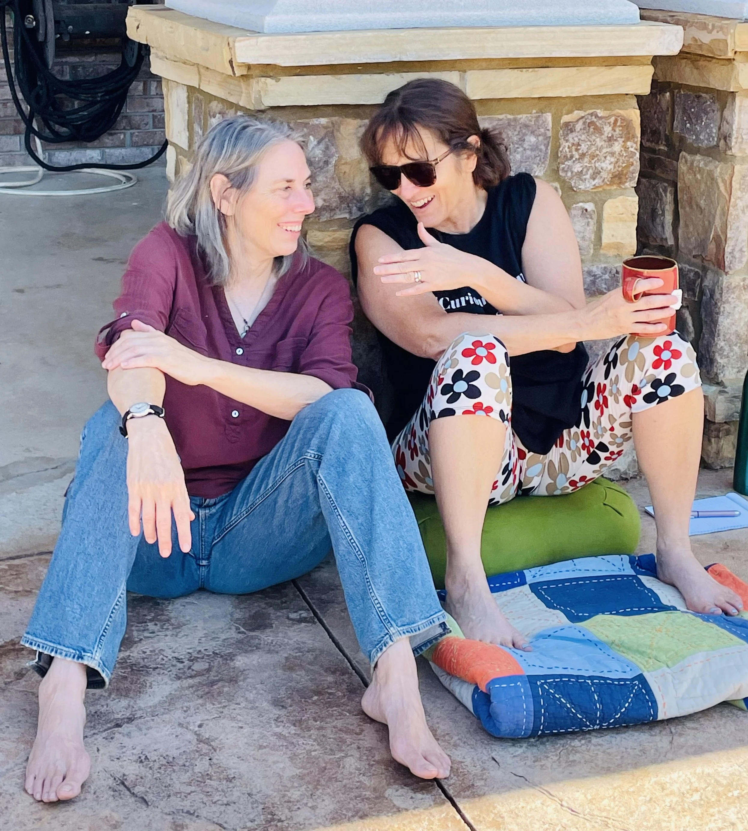 Two women sitting on a stone patio, smiling, engaging in conversation. One woman has gray hair and is wearing a maroon shirt and jeans, while the other woman, with brown hair and sunglasses, is wearing a black t-shirt and colorful floral pants. They are seated near a stone fireplace, with a red mug in the woman’s hand and a small quilted blanket on the ground.