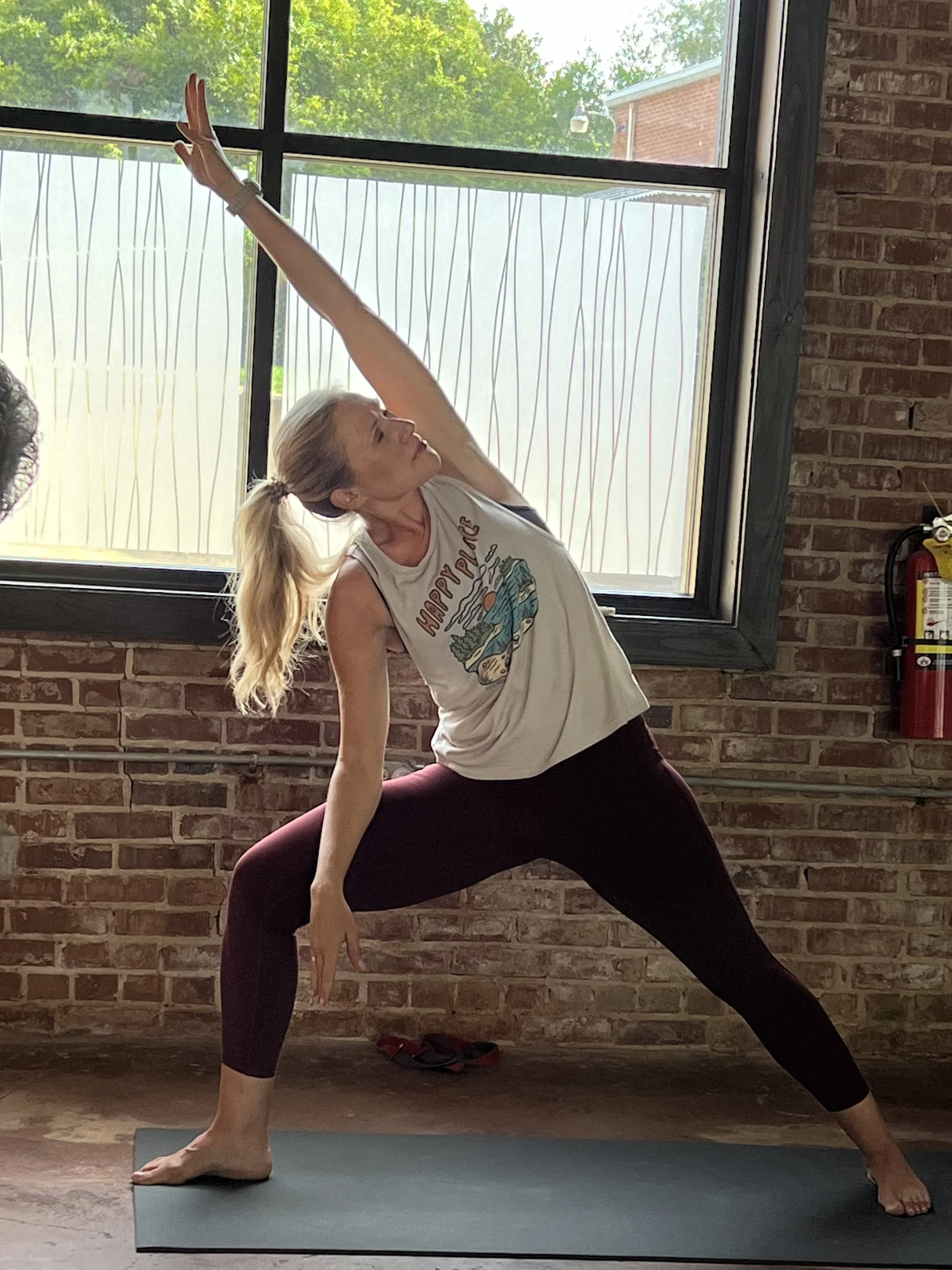 Woman practicing yoga in a lunge pose inside, with a brick wall and large window behind her.