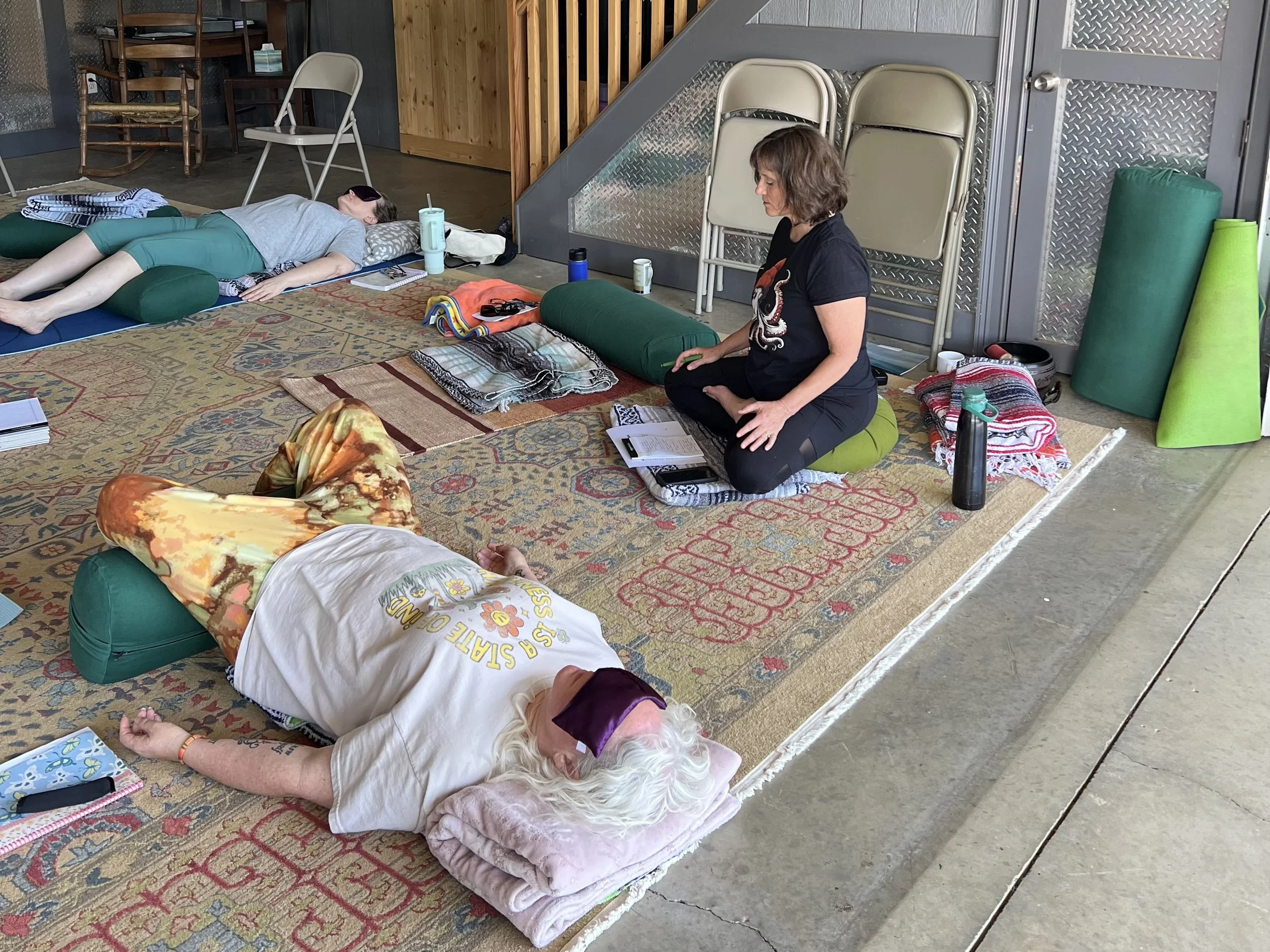 People lying on mats in a room with chairs, some with their eyes closed, participating in a relaxation or meditation session. A woman is sitting cross-legged on a cushion, with various items around her, including water bottles, a notebook, and blankets.