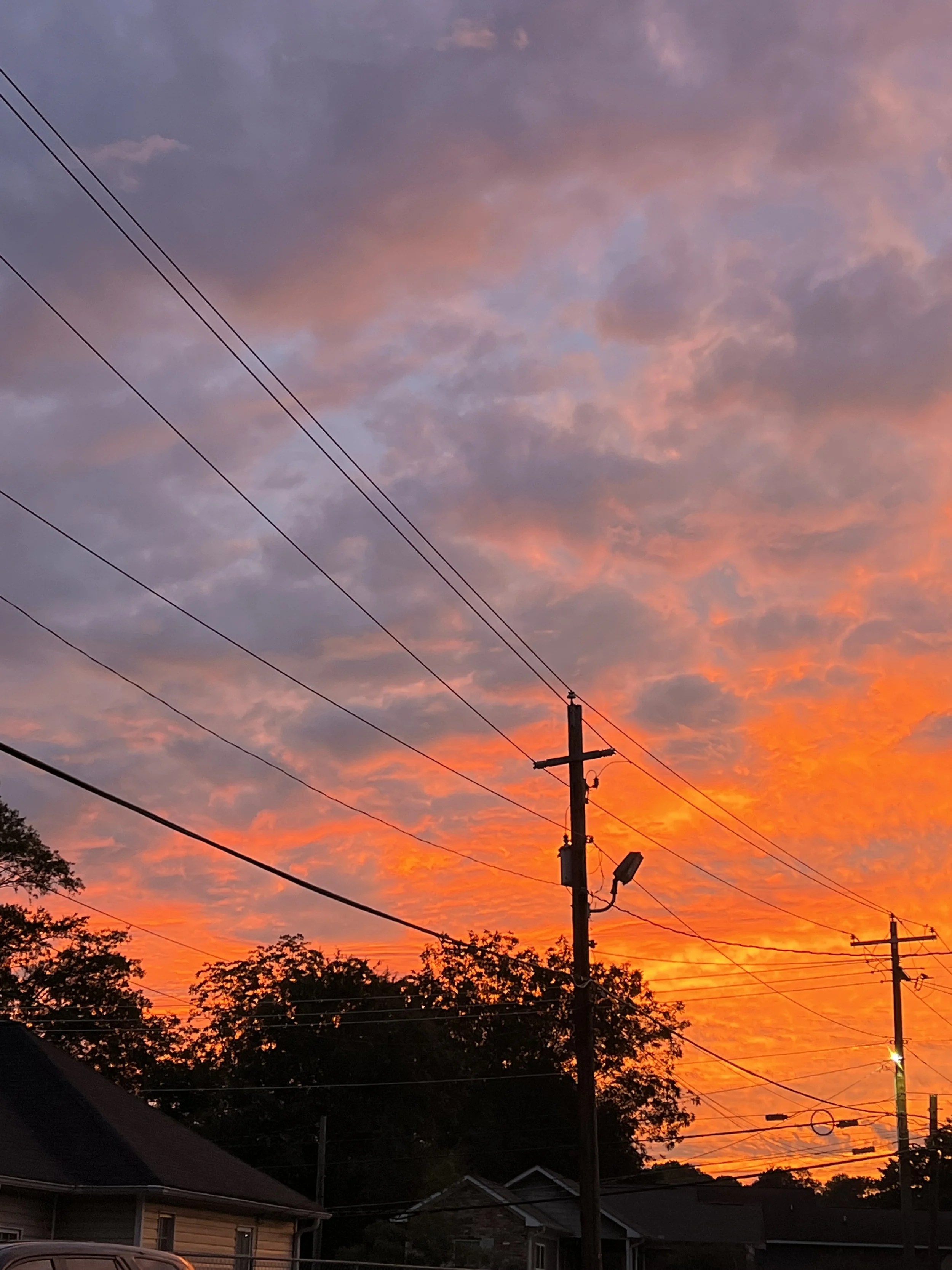 Sunset sky with colorful pink, purple, and orange clouds over rooftops and a utility pole with power lines in a suburban neighborhood.