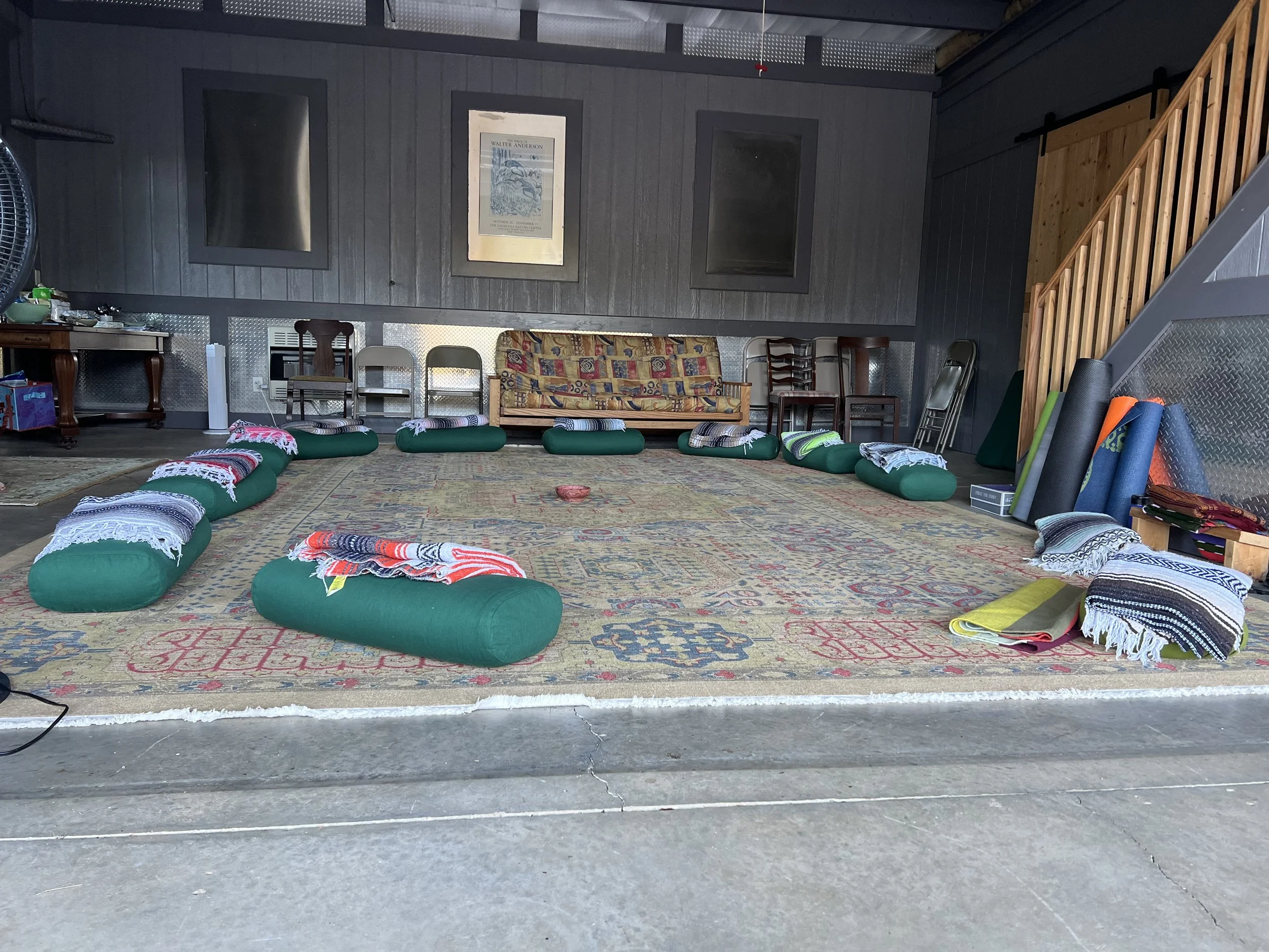 A room with a gray wall, wooden staircase, and various chairs. On the floor, there are green cushions with towels, and a colorful area rug. Items are scattered on a small wooden table to the left, and three framed pictures or posters hang on the wall.